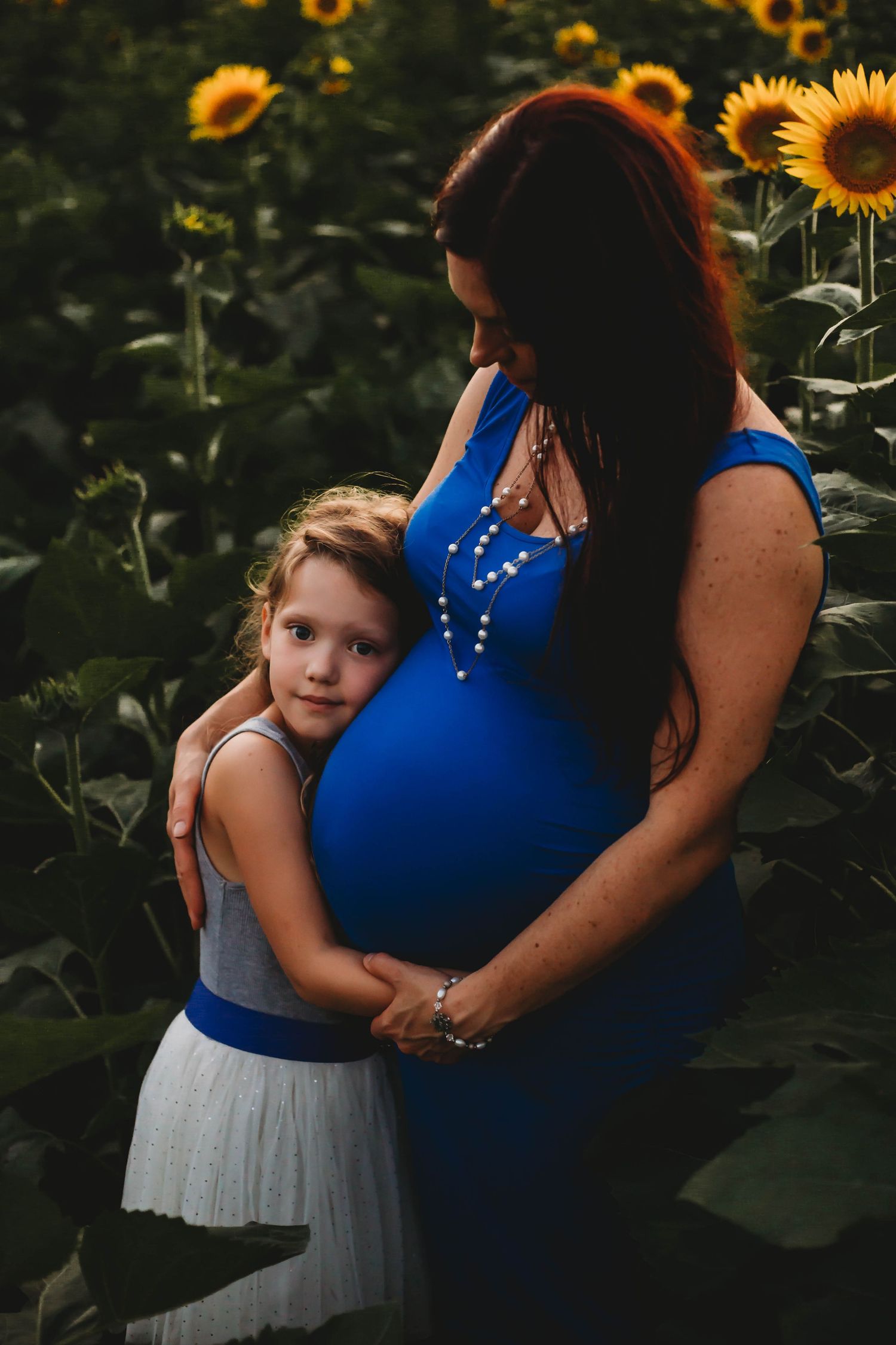 Young girl hugging pregnant woman in blue dress surrounded by sunflowers at dusk.