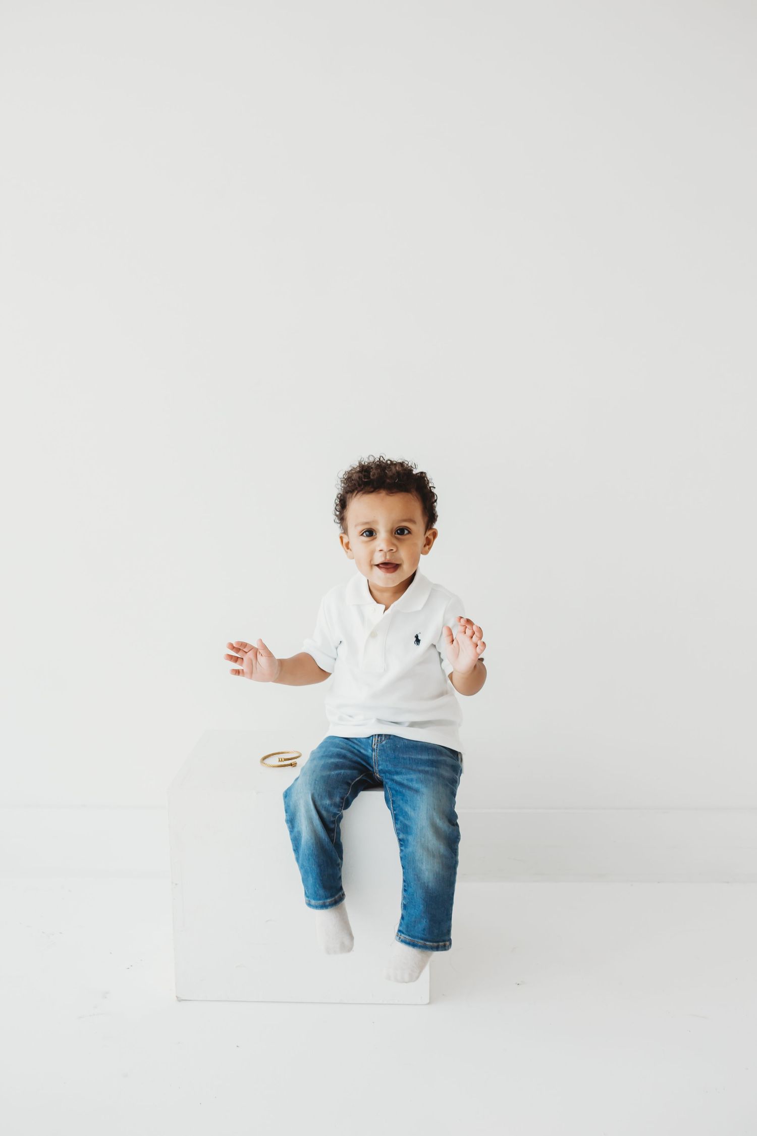 Happy toddler boy in white polo and jeans sitting on a white cube, arms raised.