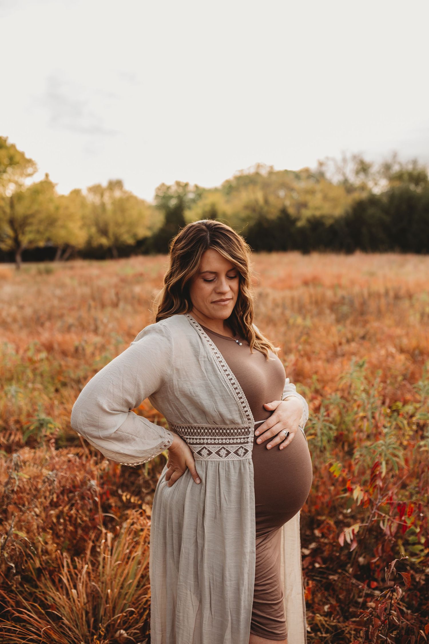 Pregnant woman in bohemian white cardigan cradling her baby bump in an autumn field at golden hour.
