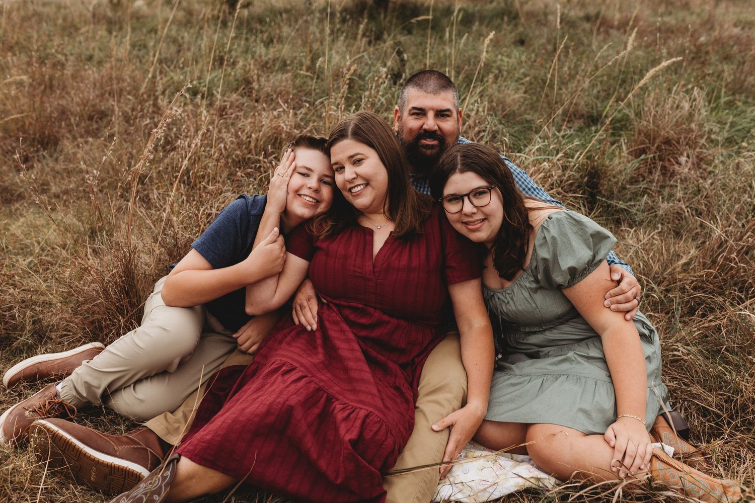 Family of four sits closely together smiling in a golden dry grass field.