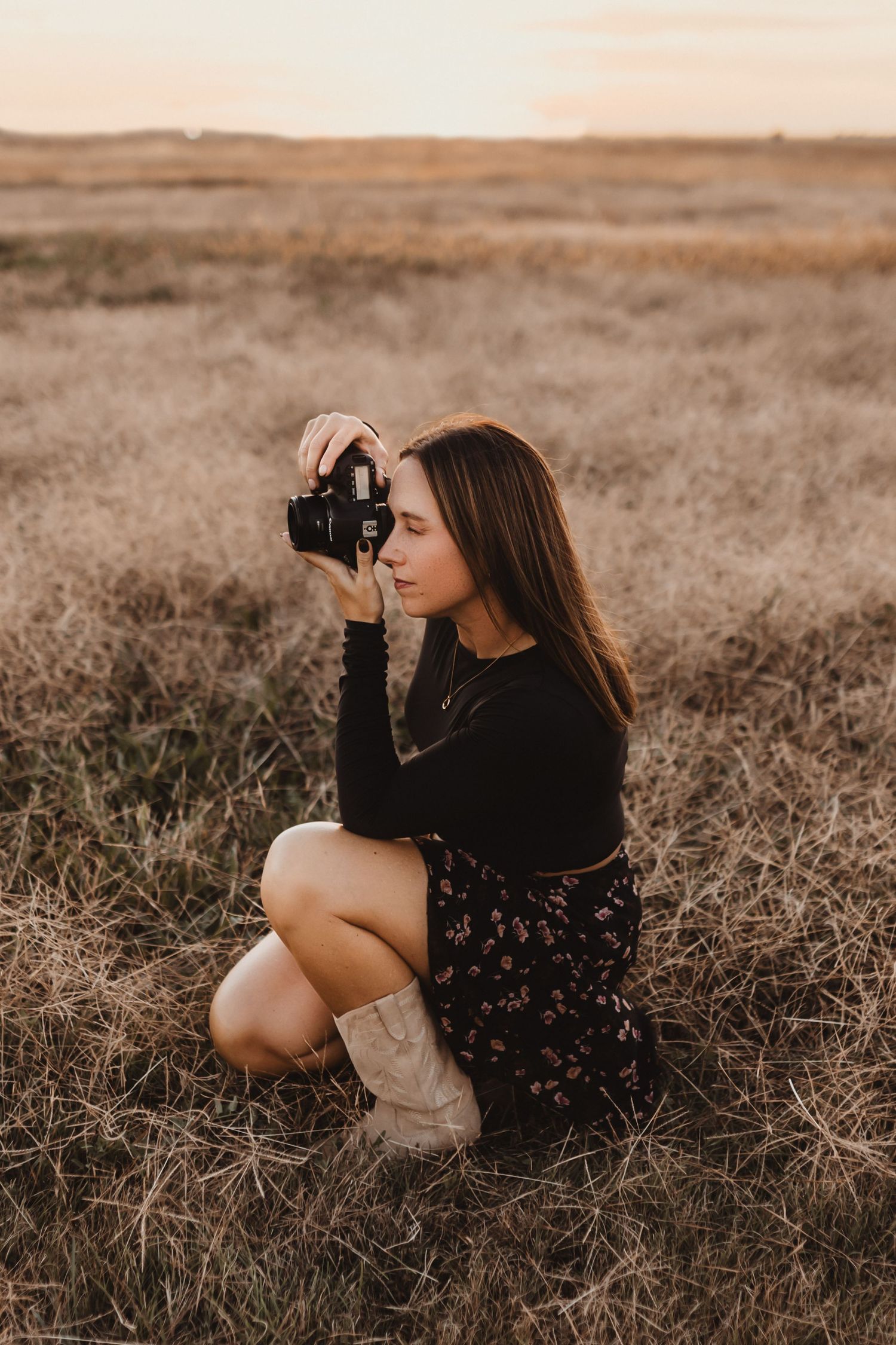 Young woman crouching in golden field at sunset holding a camera.