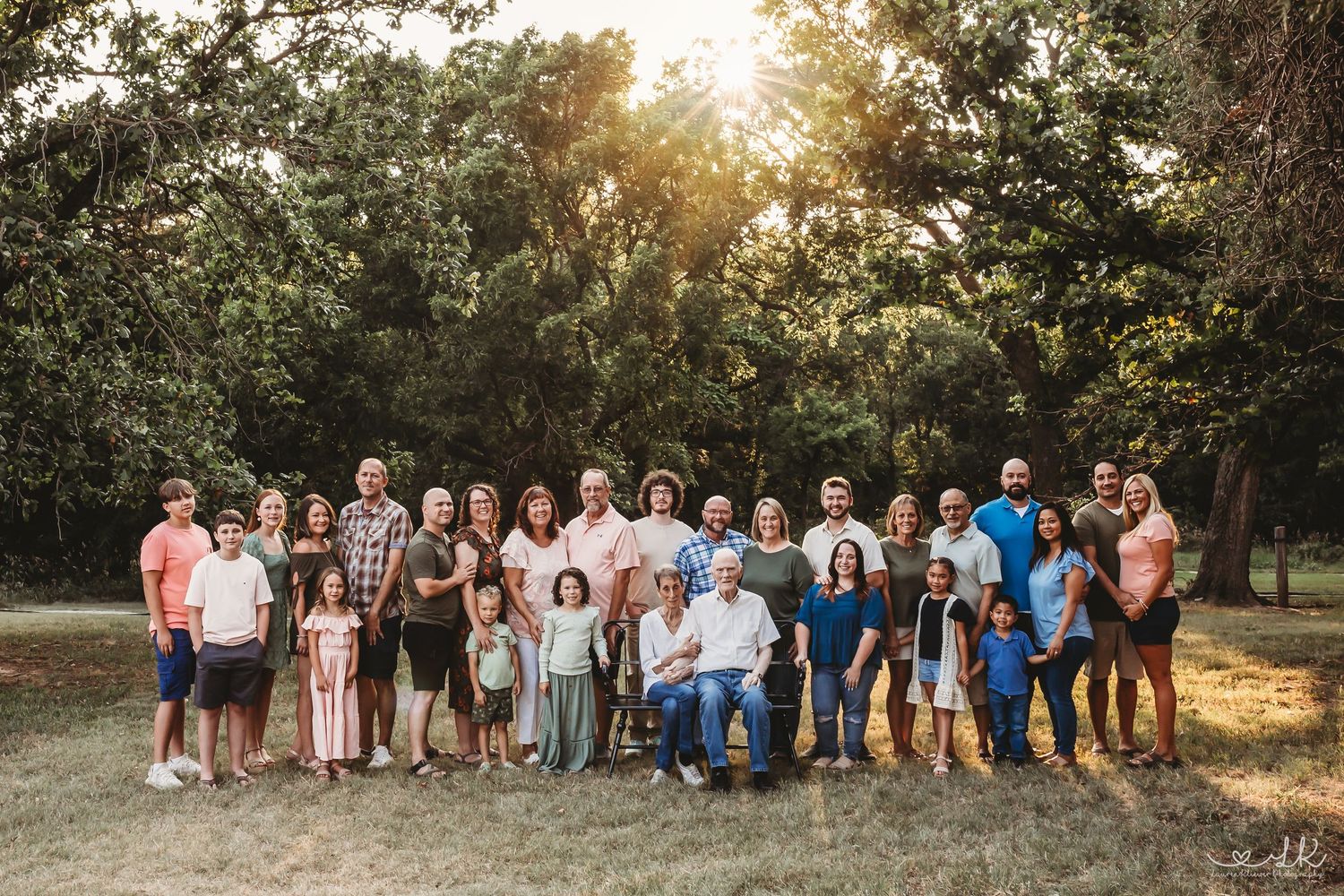 Large extended family group portrait outdoors under sunlit trees in a park setting.