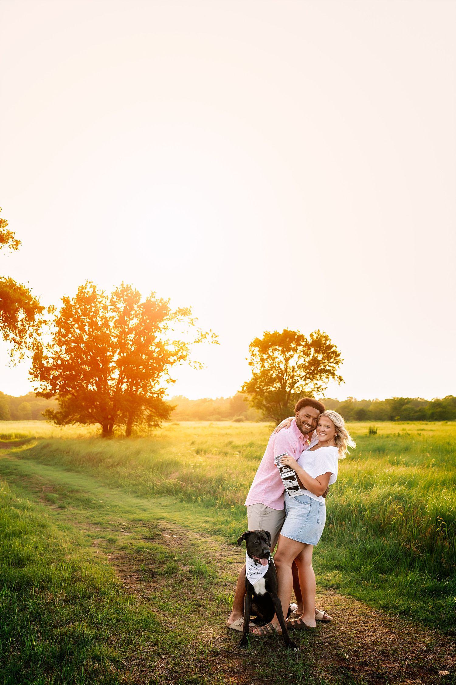 Couple embracing at golden sunset on a rural path with their black dog wearing a bandana in a green field.