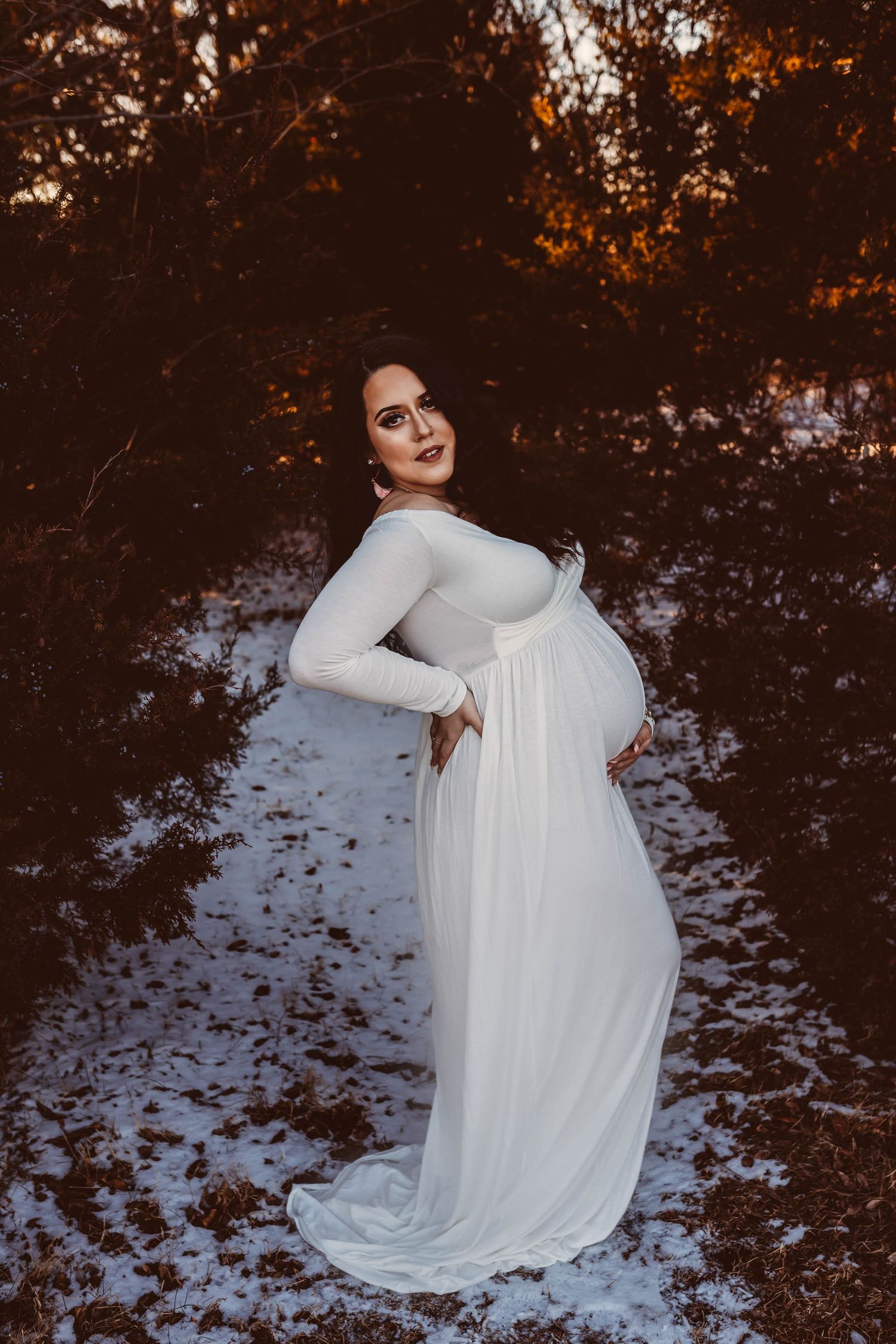 Pregnant woman in white gown poses gracefully in snowy winter forest during golden hour.