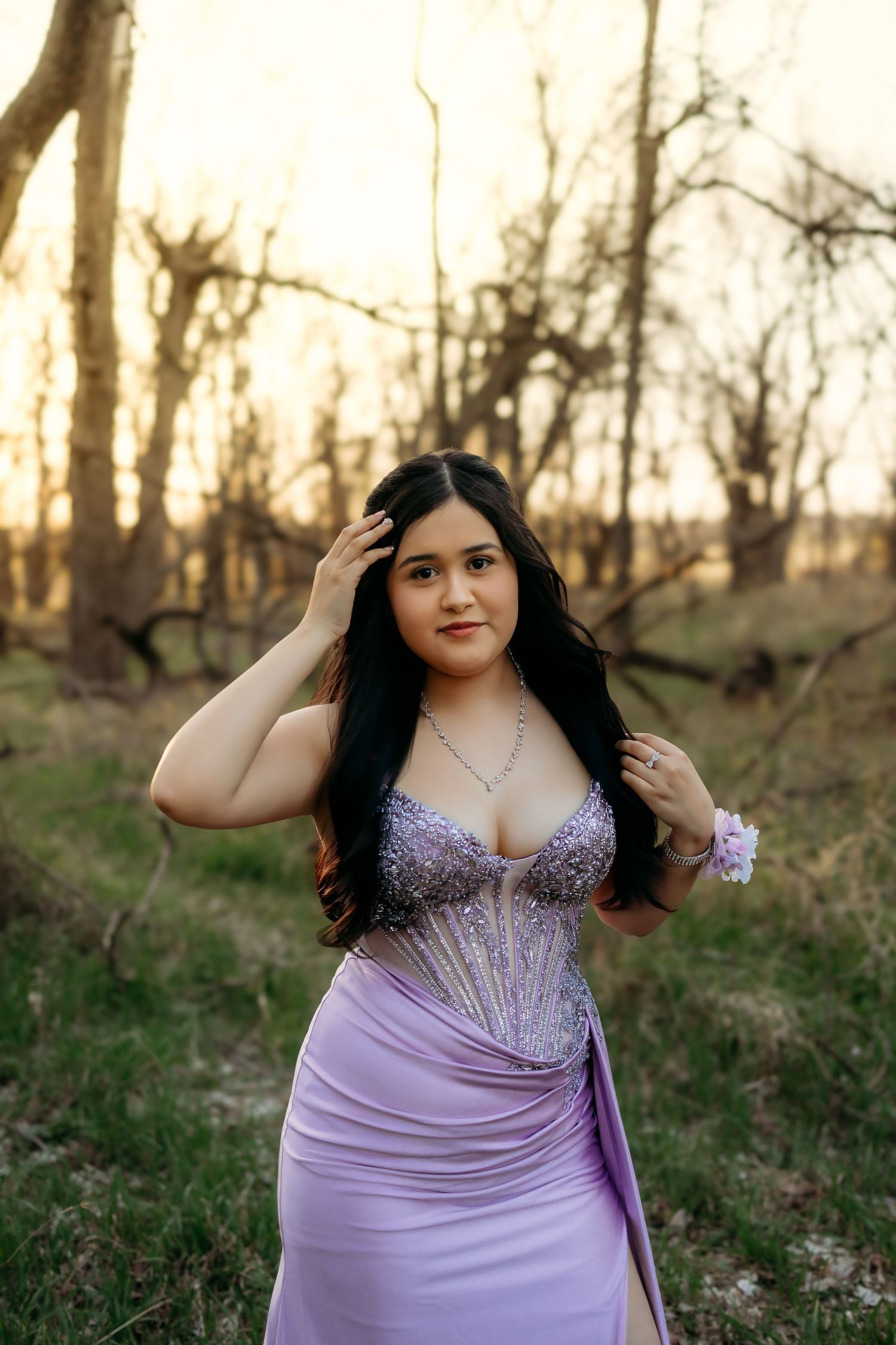 A young woman in a sparkling lavender prom gown poses outdoors among bare trees at sunset.