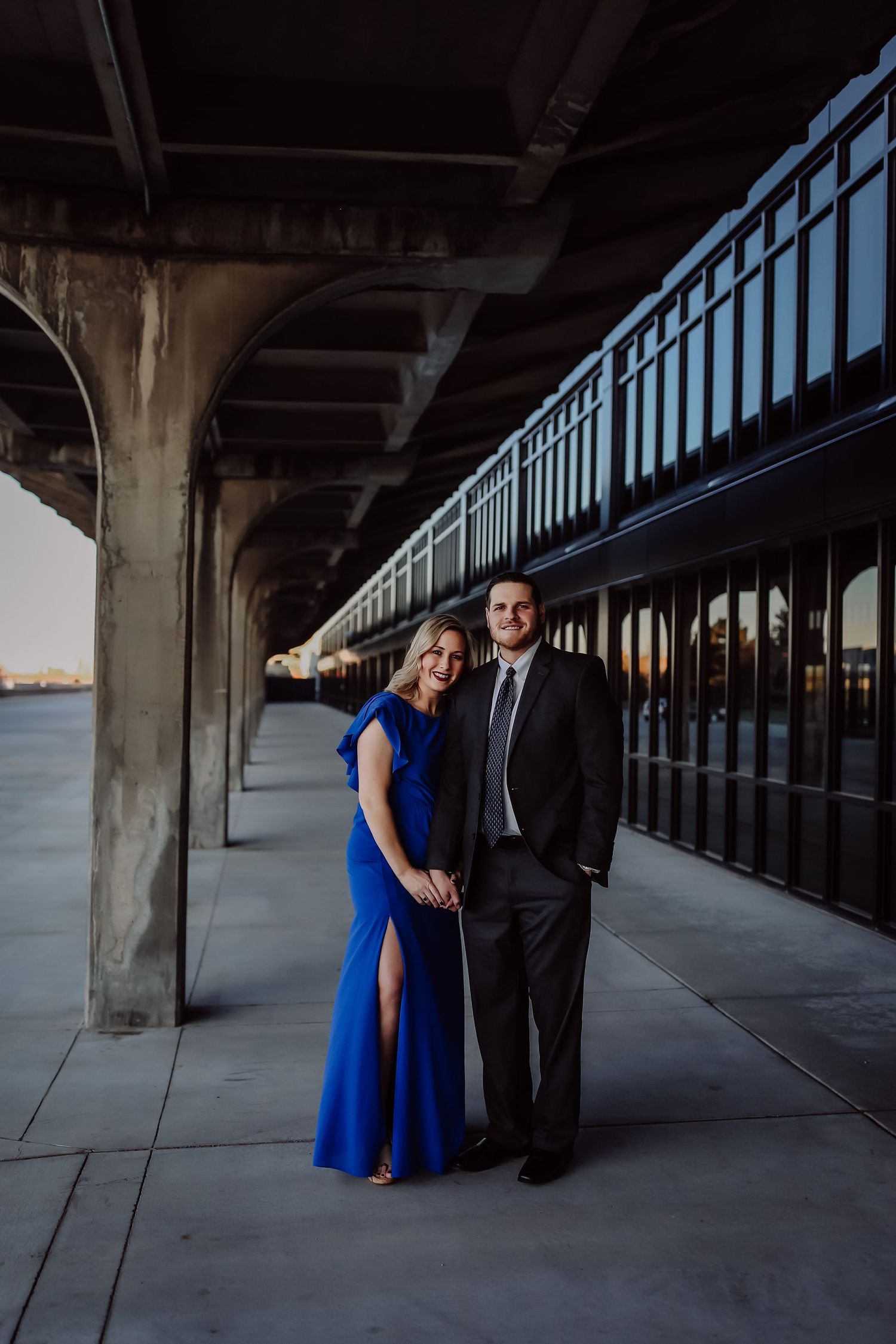 Elegant couple in formal attire posing under a bridge structure at sunset near waterfront.