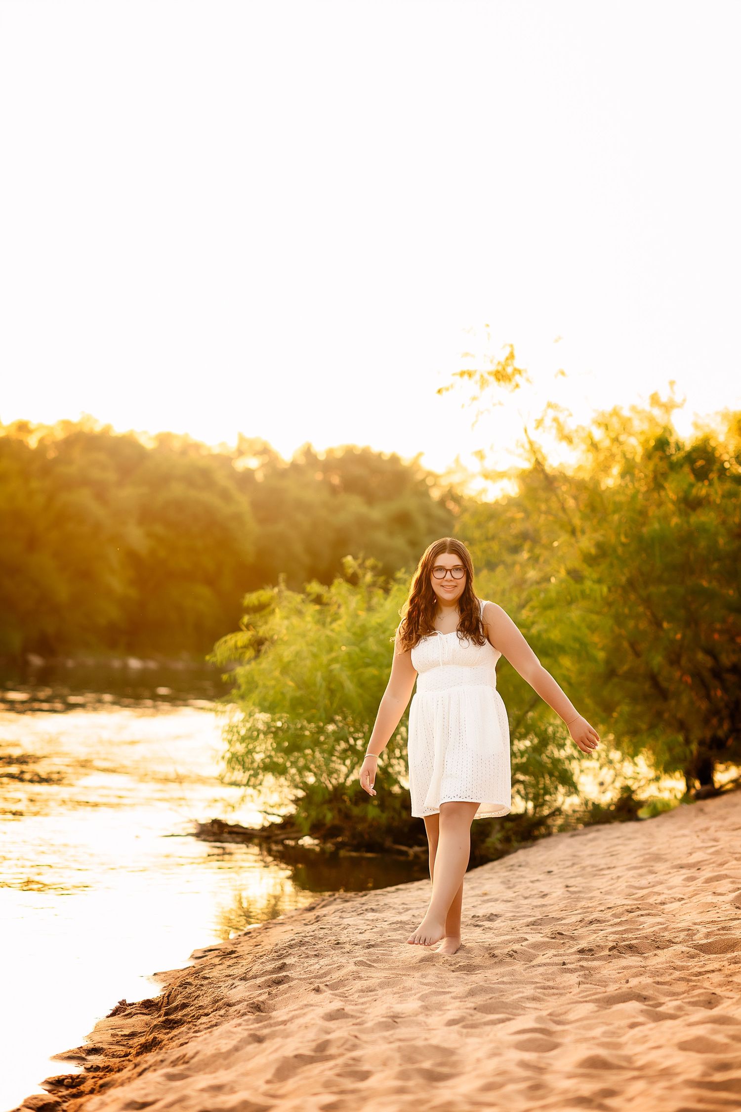 Young woman in white dress walking barefoot on sandy beach at golden sunset near lush green trees.