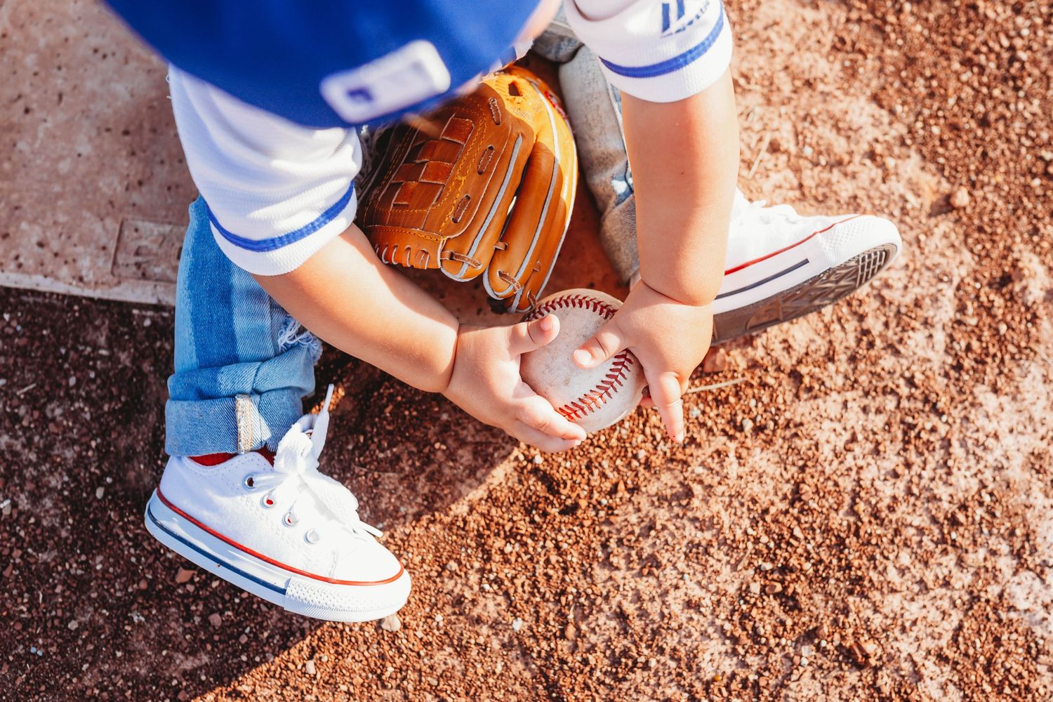 Young baseball player picking up a ball wearing a mitt, white sneakers, and blue uniform on a dirt field.