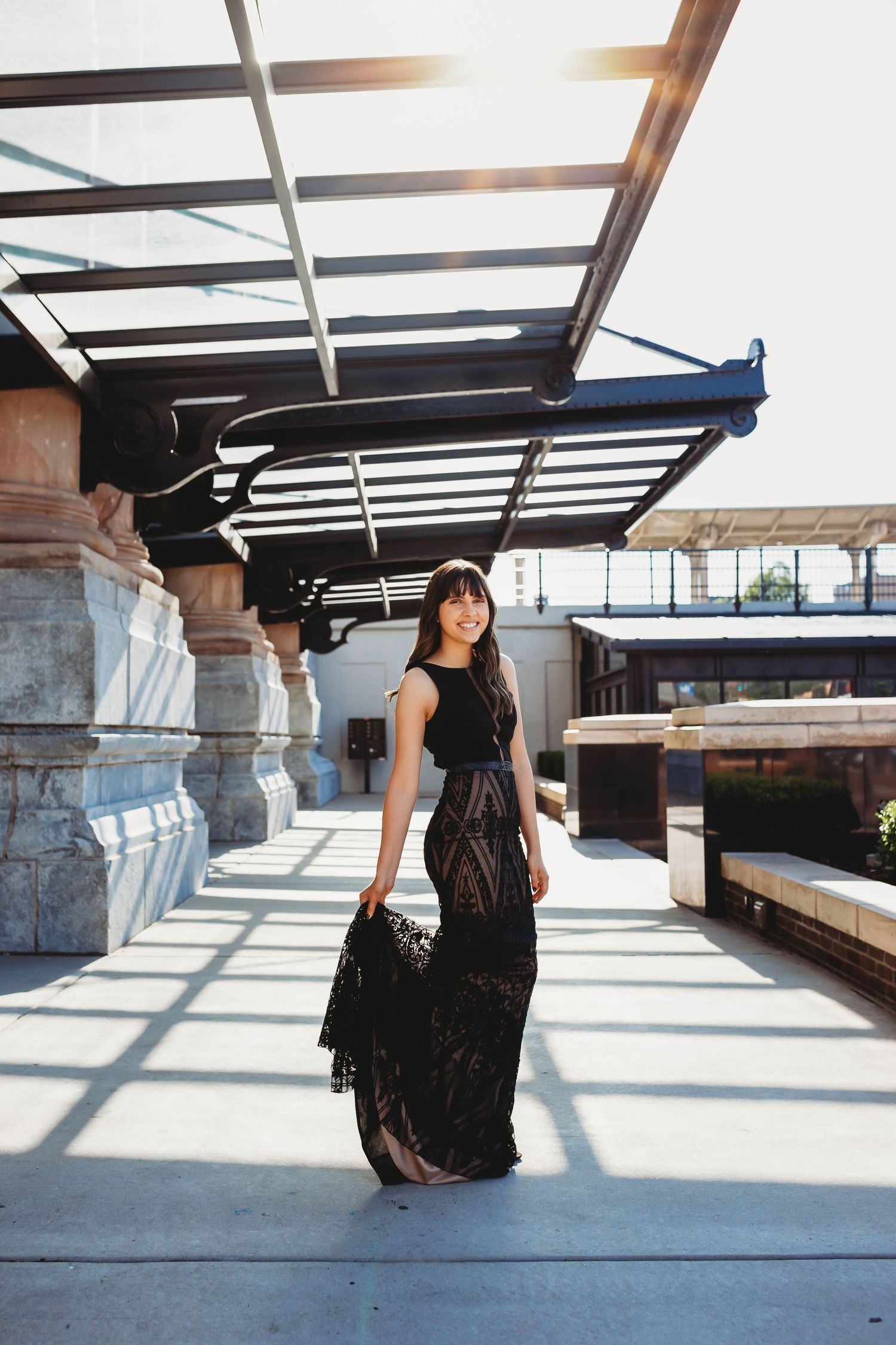 Elegant woman in black lace gown walking on sunlit rooftop terrace with columns.