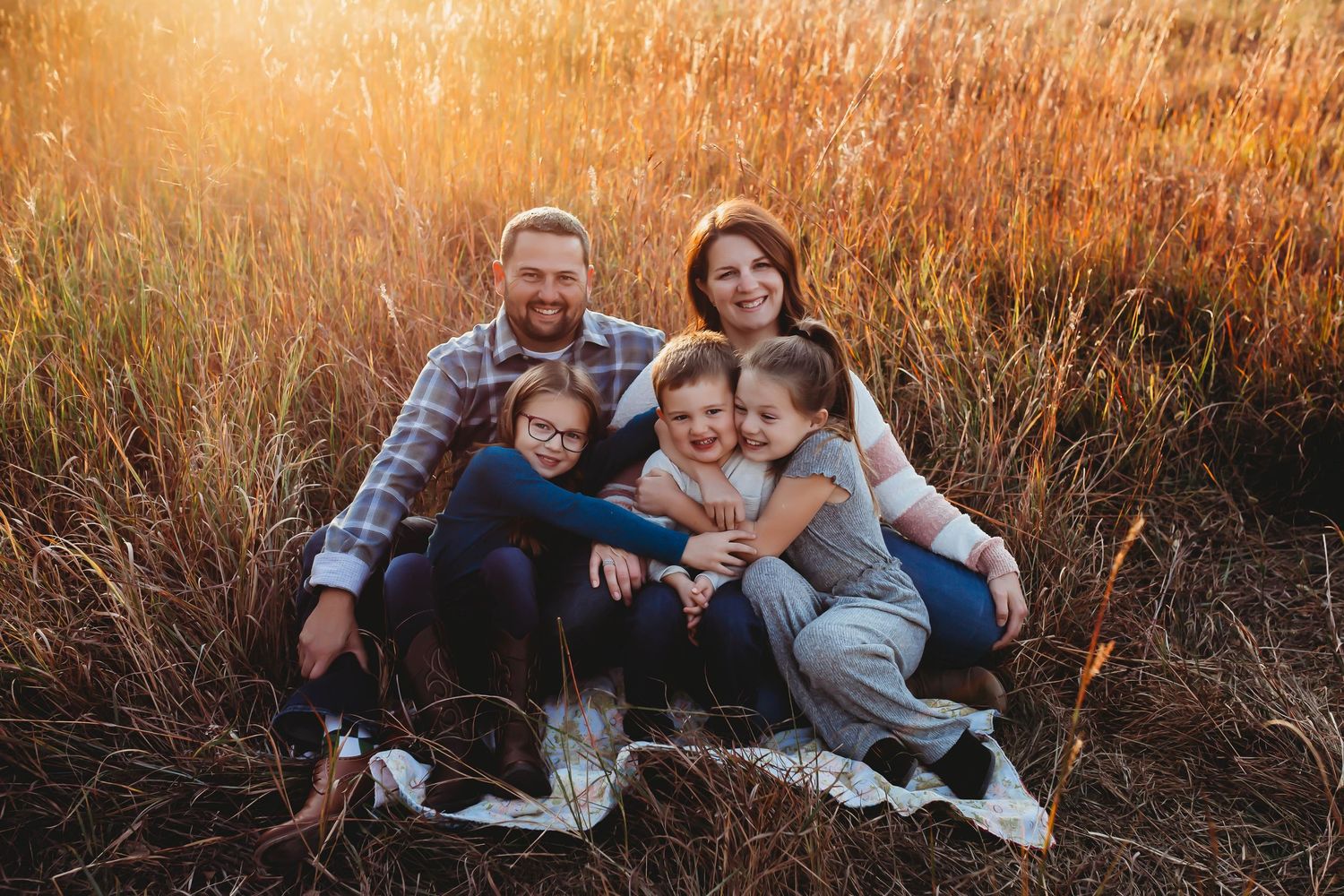 Happy family of five sitting together on blanket in golden sunlit tall grass field.