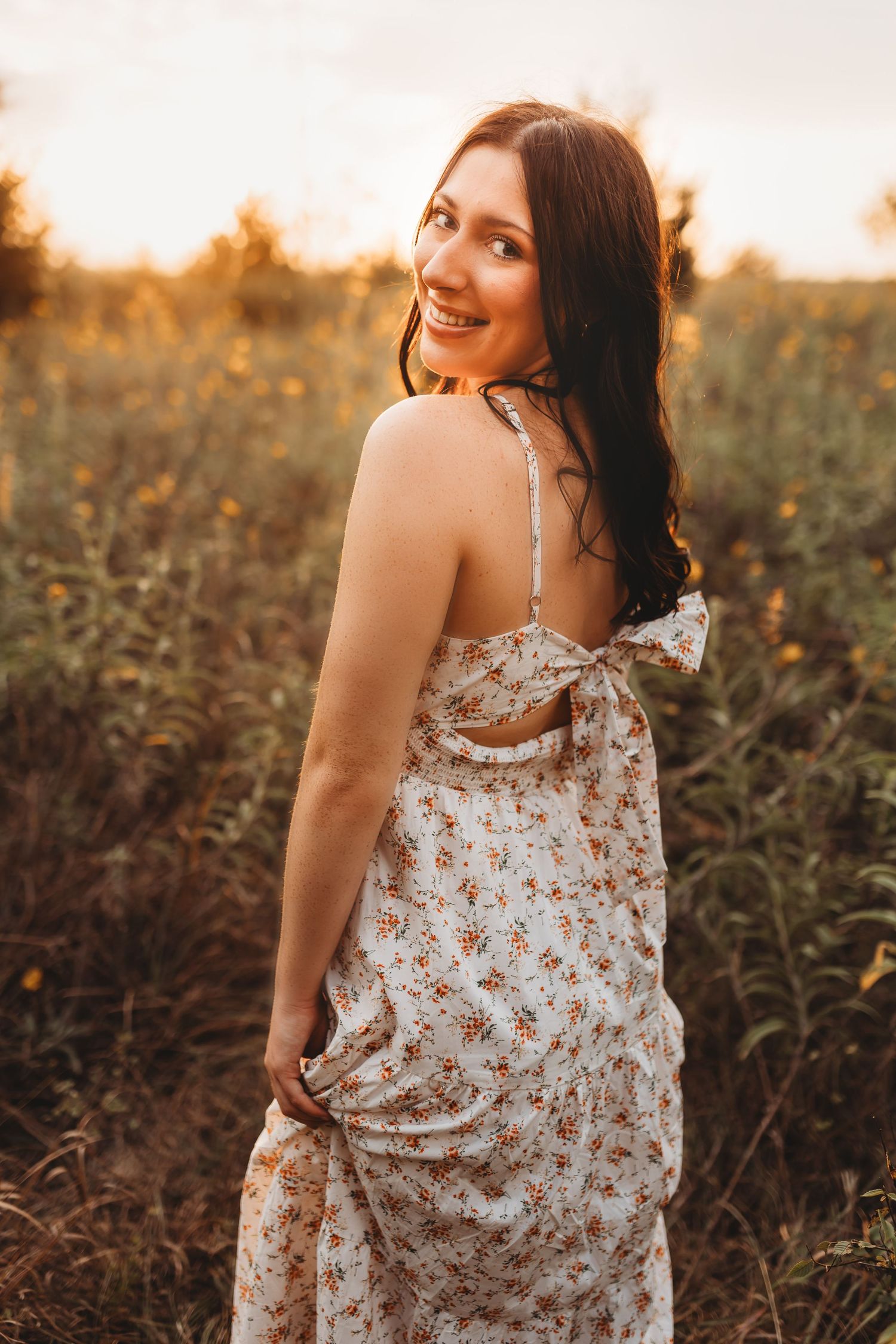 Young woman in floral sundress smiling over shoulder at golden hour in field.