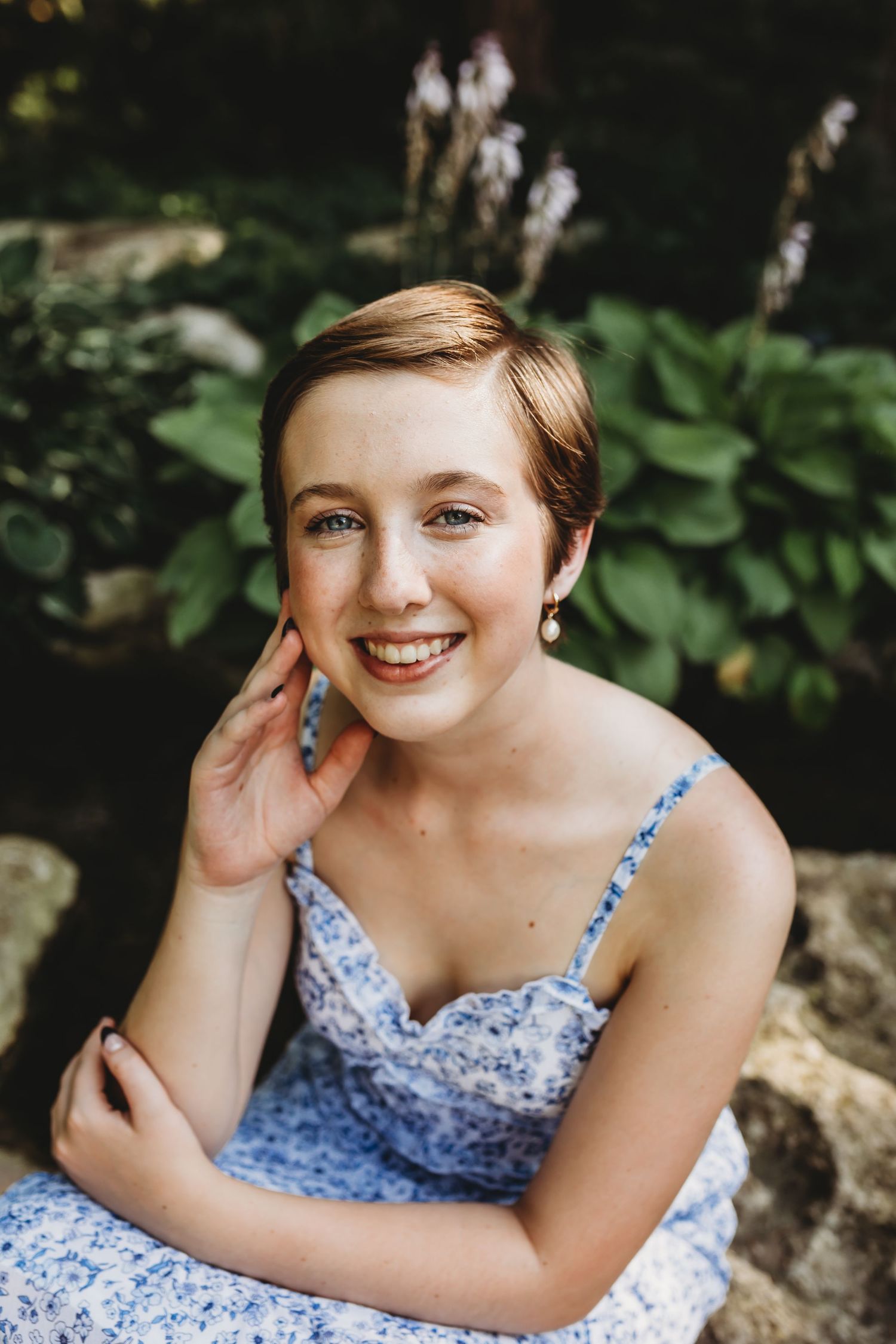 A smiling teenage girl in a blue floral dress poses gracefully among lush green garden foliage.