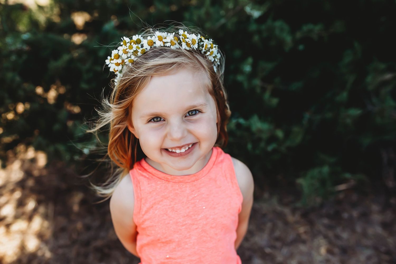 Smiling young girl wearing a daisy flower crown and coral sleeveless top outdoors.