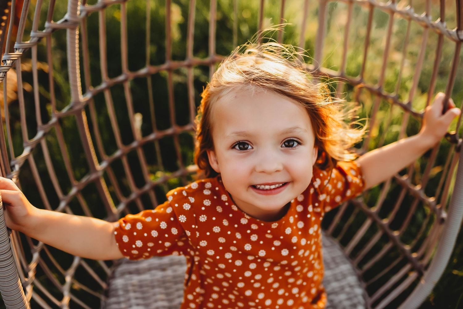 Adorable little girl in a red polka dot dress smiling while sitting in a wicker hanging chair.