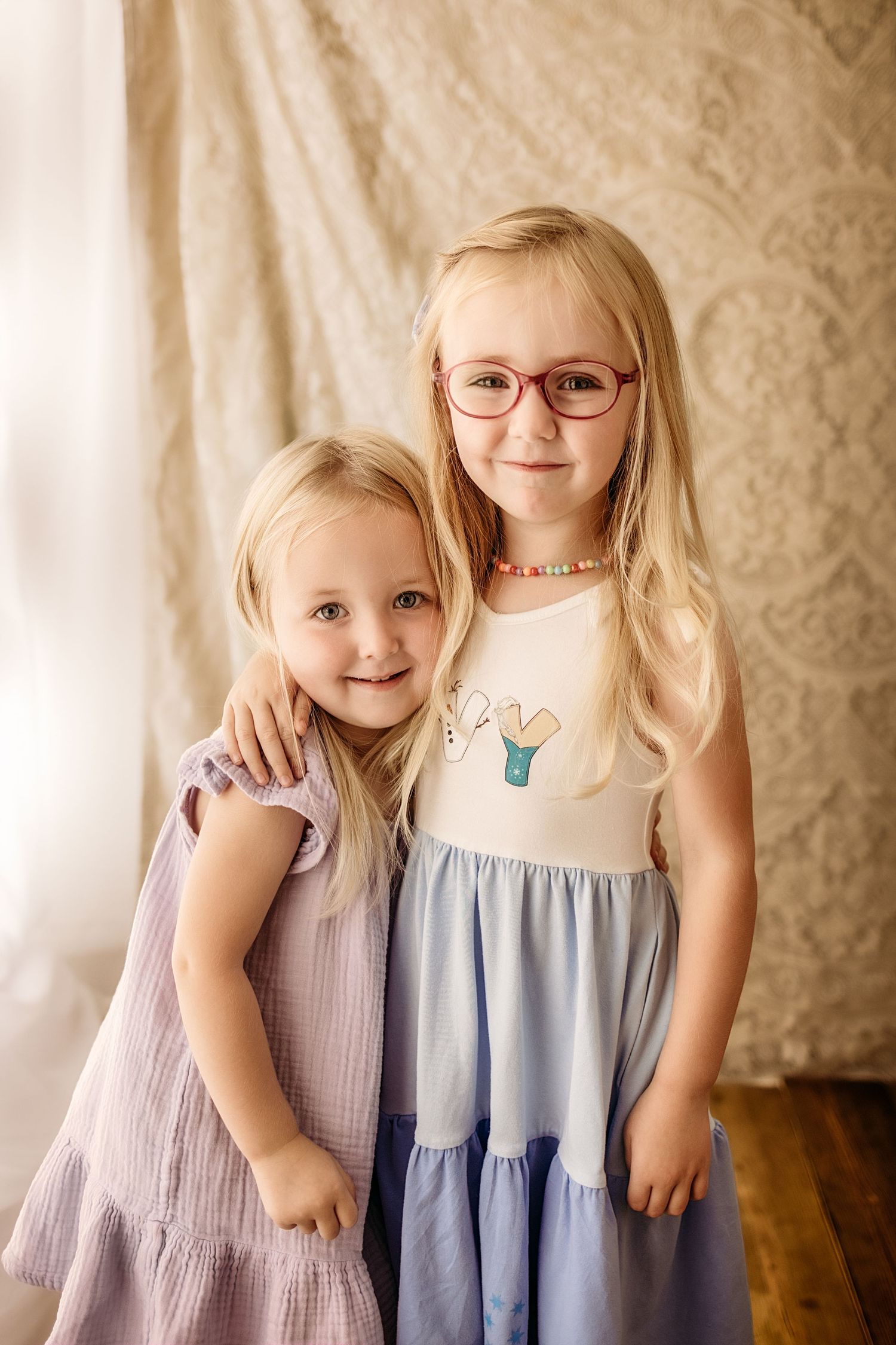 Two young blonde sisters posing together, one wearing glasses and a blue dress, the other in a lavender dress.