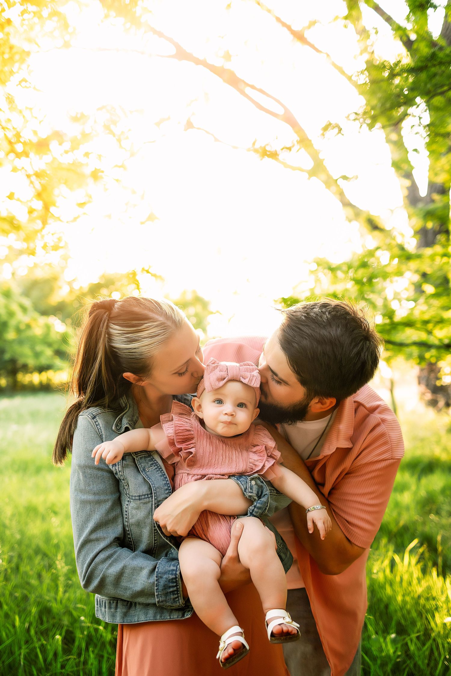 Parents kissing their baby girl outdoors in golden sunlight, family portrait in a lush green park setting.