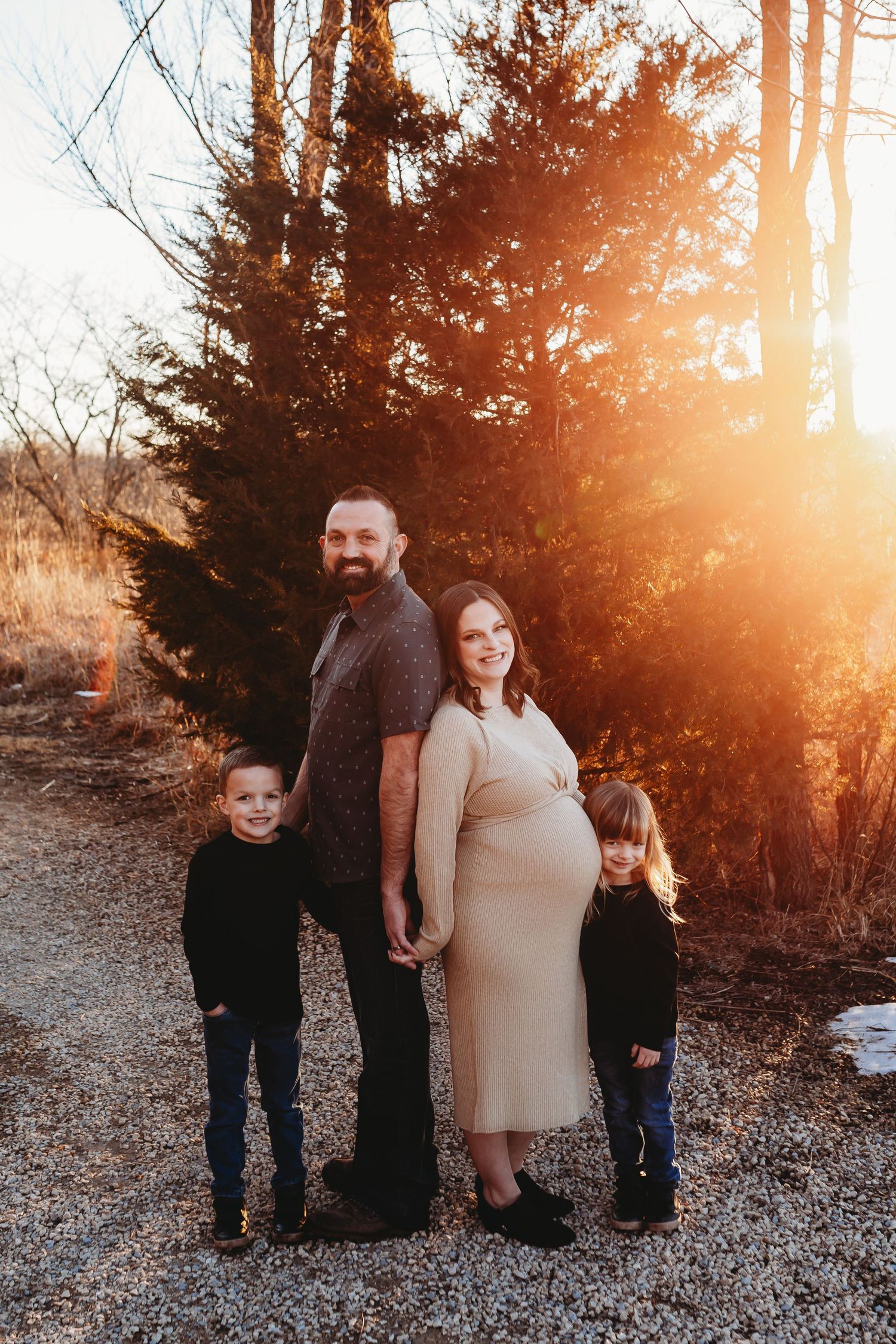 Pregnant family of four posing at golden hour near evergreen trees on a gravel path.