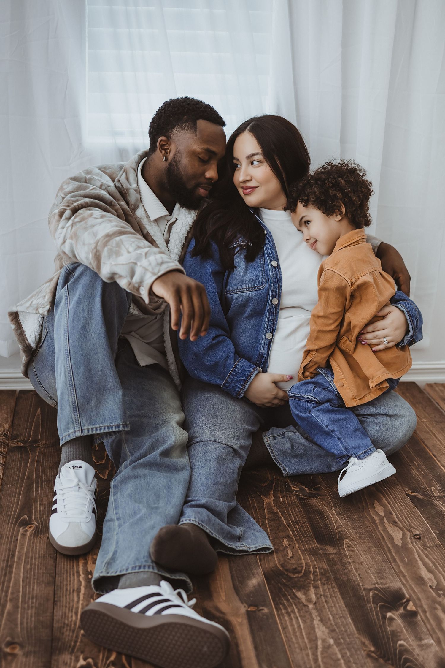 A multiracial family sitting on hardwood floor, pregnant mother in denim, father and curly-haired toddler embracing warmly.