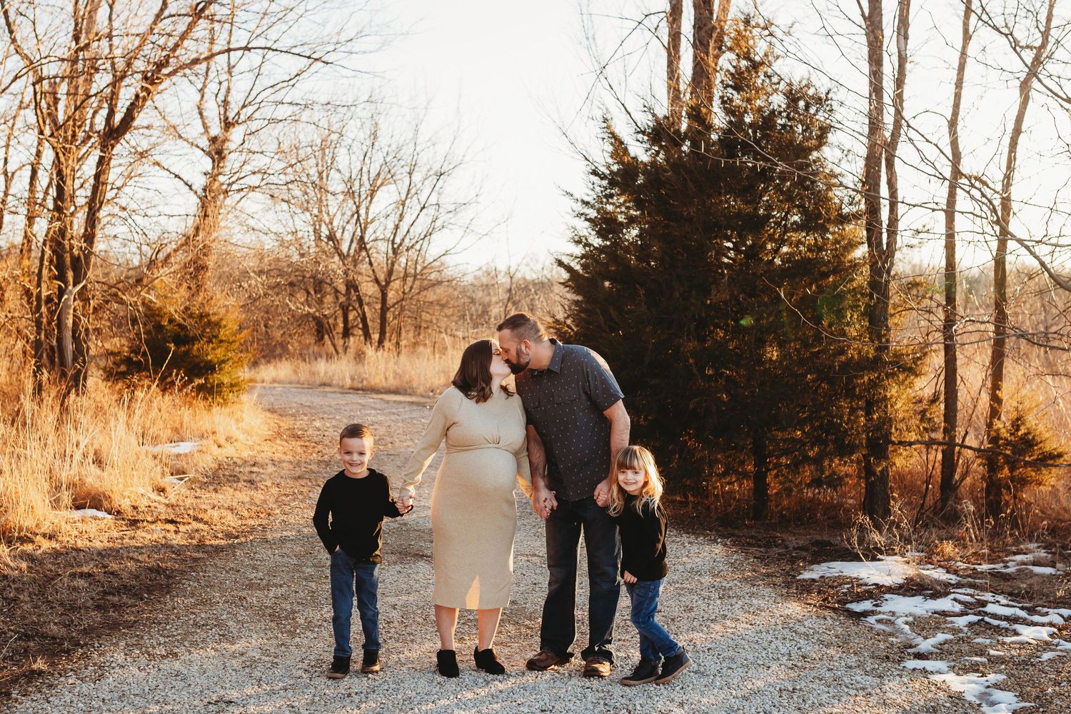 Family of four sharing a kiss on a gravel path surrounded by winter trees.