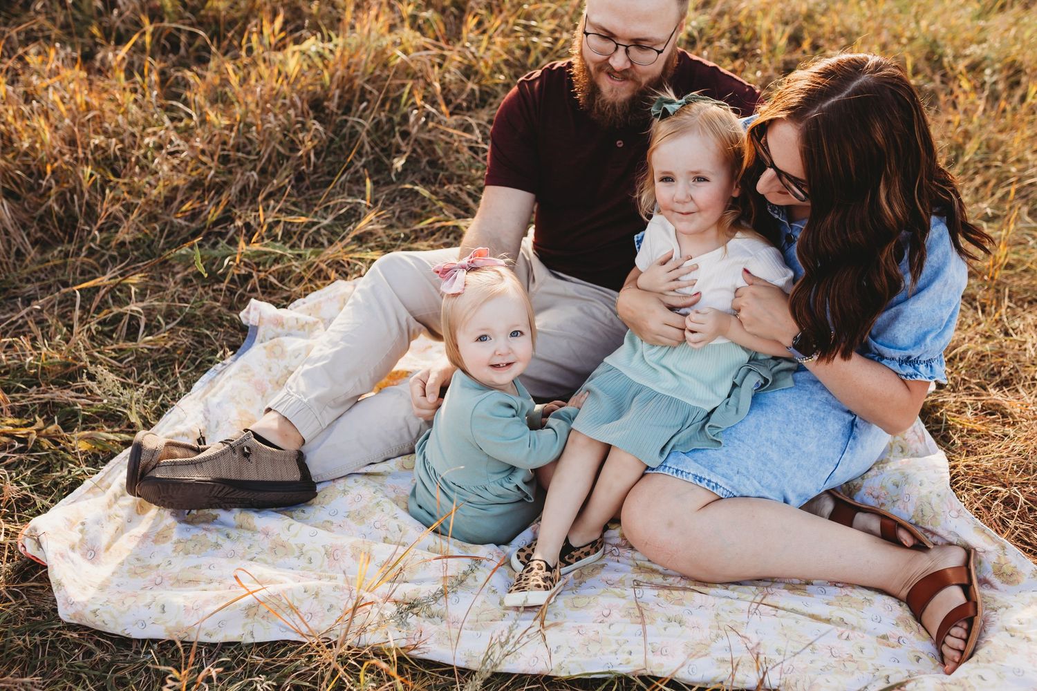 Young family sitting on a blanket in a golden field with two toddler girls.