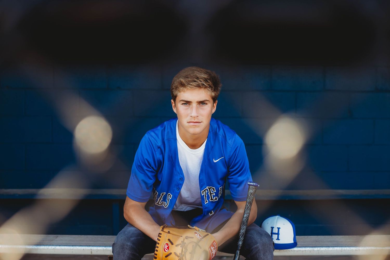 Teen baseball player in blue jersey sitting in dugout holding glove for senior portrait photo.