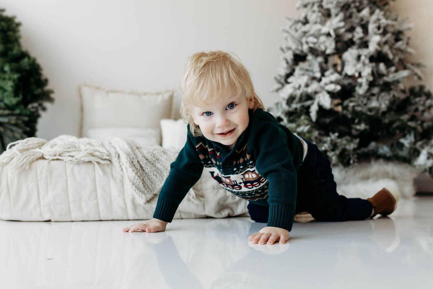 A cheerful blonde toddler in a festive sweater crawls on a white floor near a Christmas tree.