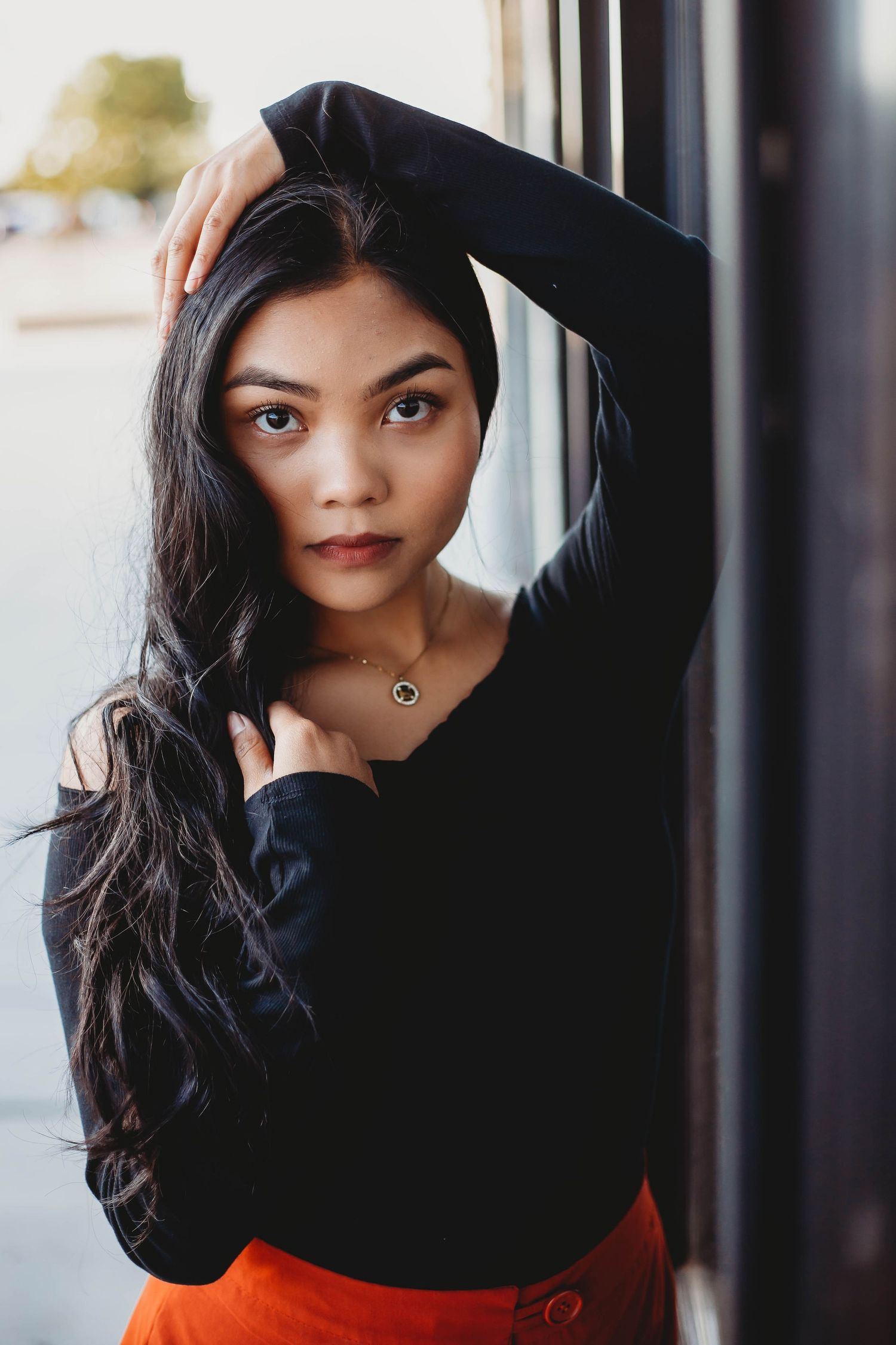 A young woman with long dark hair in a black top and orange skirt poses by a window.