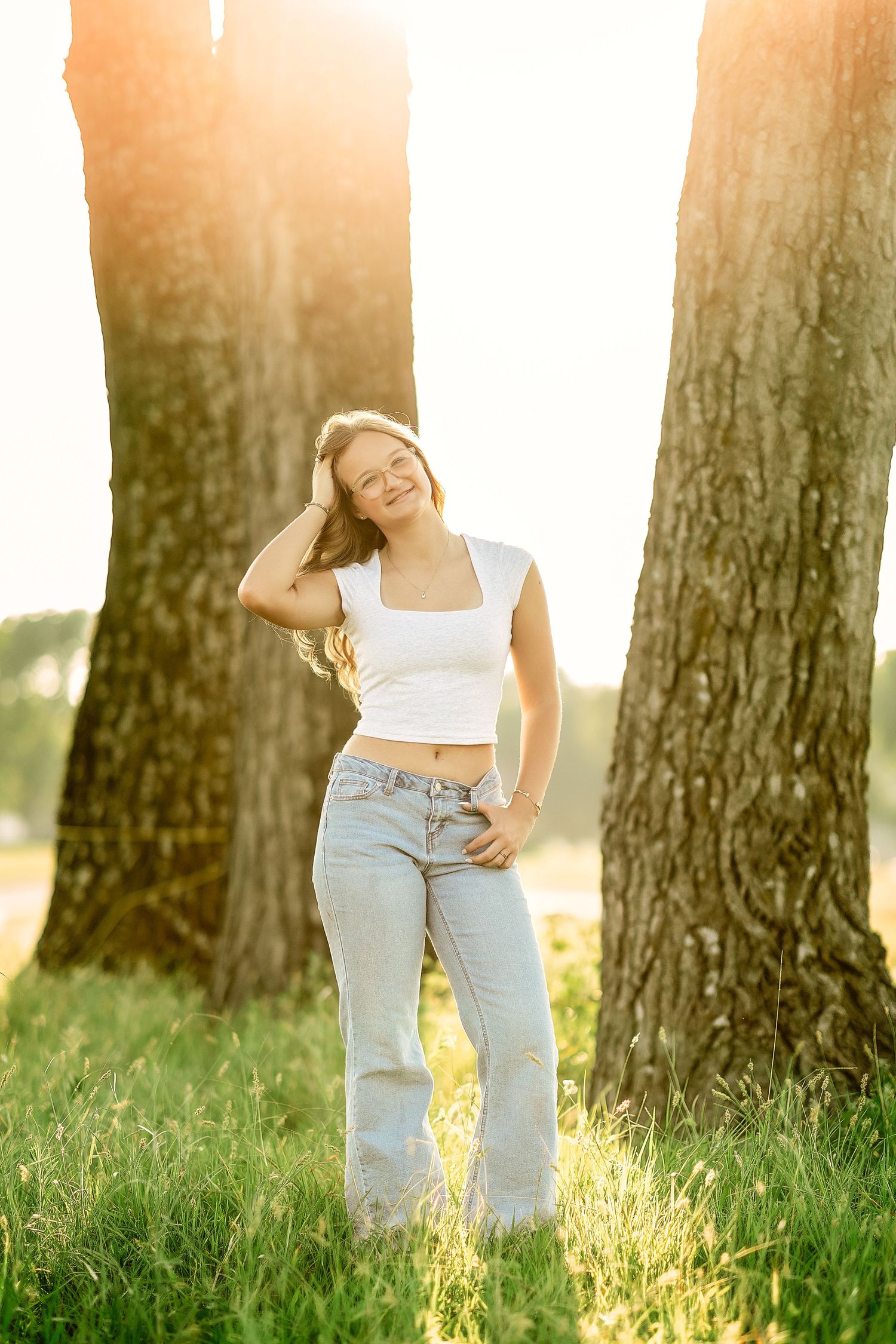 Young woman in white crop top and jeans smiling between two trees in golden sunlight outdoors.