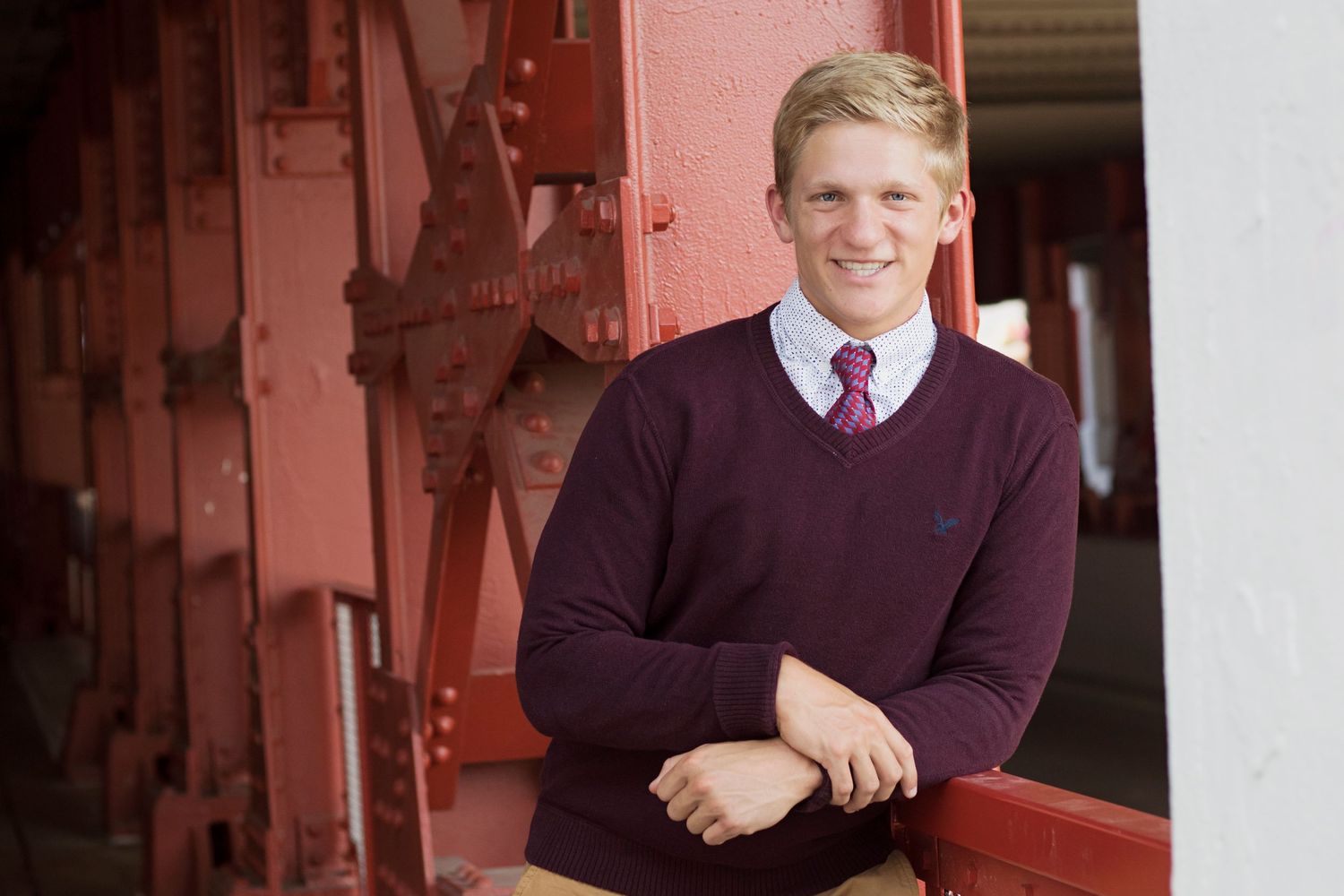 Young man in purple sweater and tie smiling outdoors near red metal structure in a professional portrait.