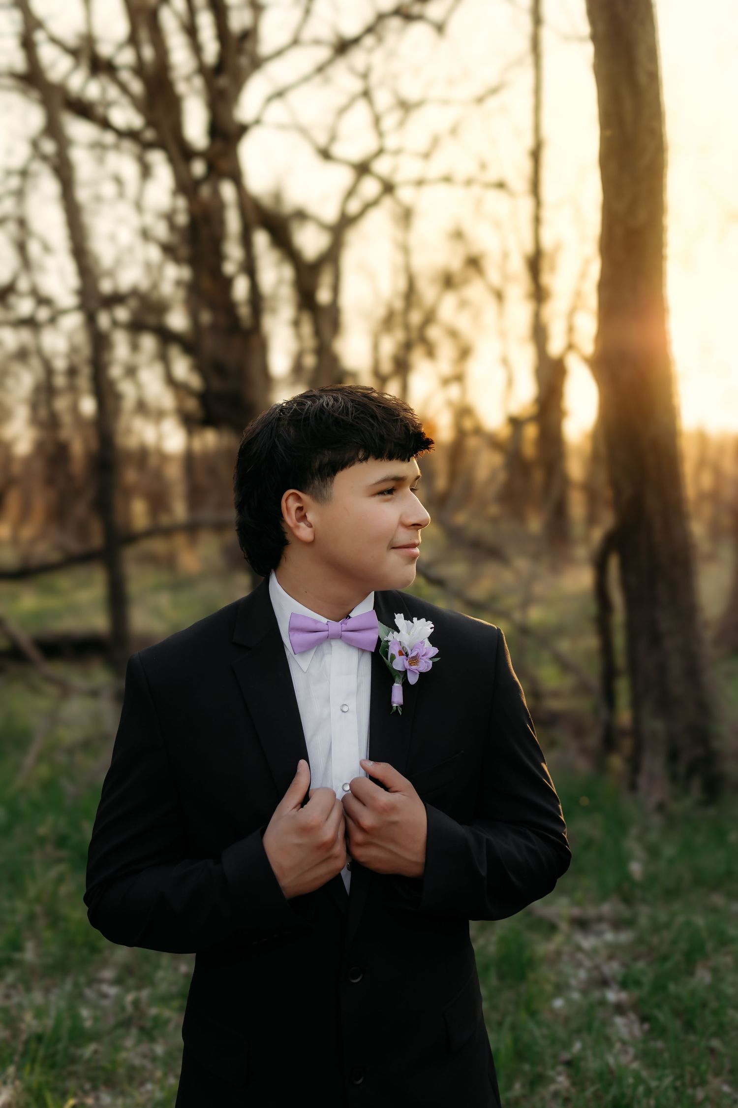 A young man in a black tuxedo with purple bow tie and boutonniere poses at sunset outdoors.