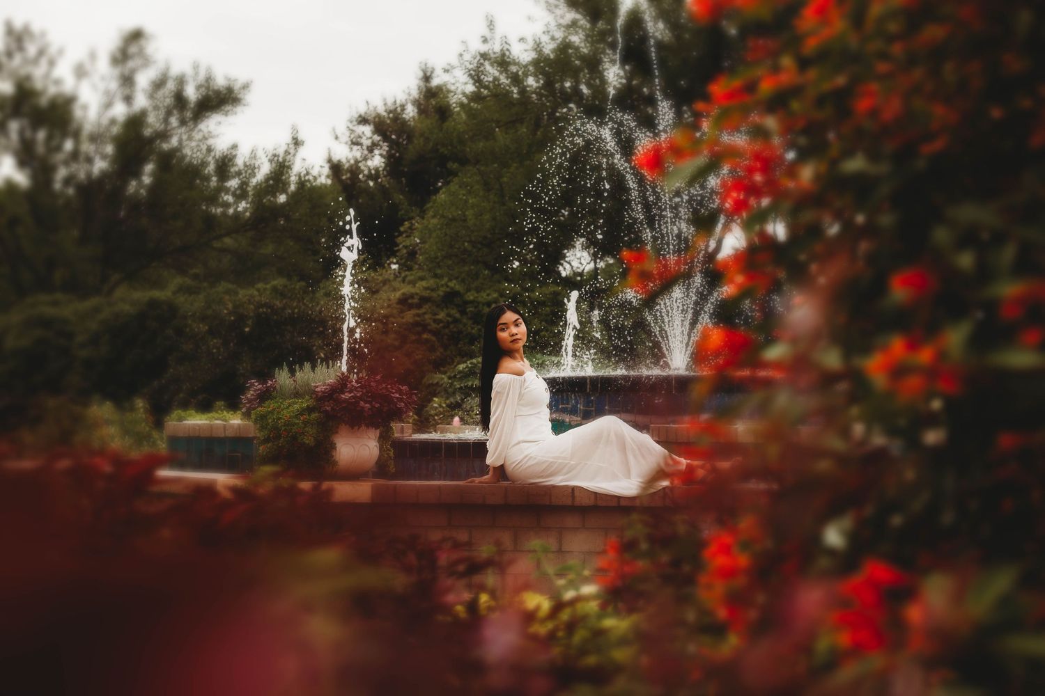 Woman in white dress sitting by a fountain surrounded by red flowers in a garden.