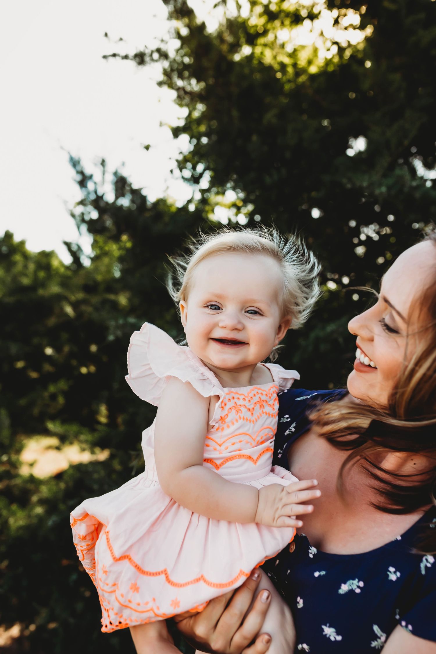 Smiling red-haired mother holding laughing blonde baby girl in pink dress outdoors in sunlight.