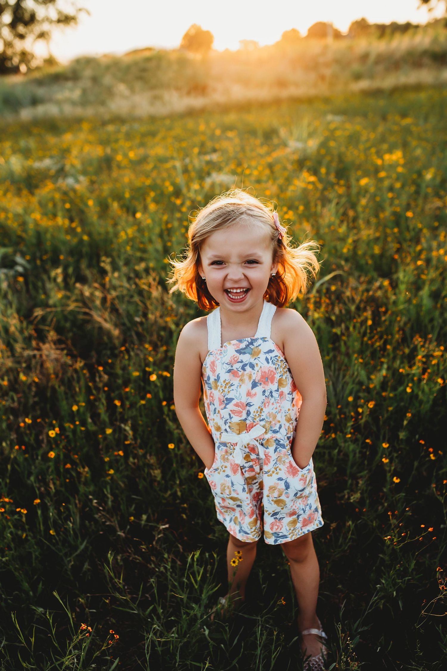 Joyful little girl in floral romper laughing in a sunny wildflower field at golden hour.