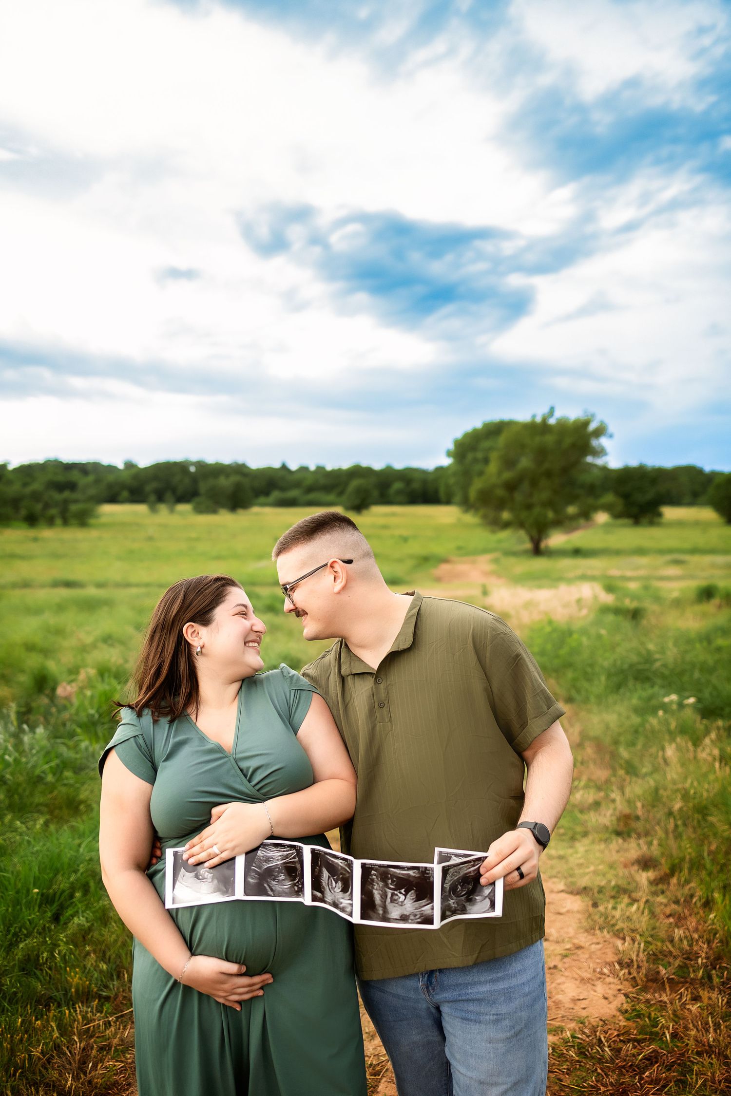 Smiling pregnant couple holding ultrasound photos in a green field, celebrating their pregnancy outdoors.