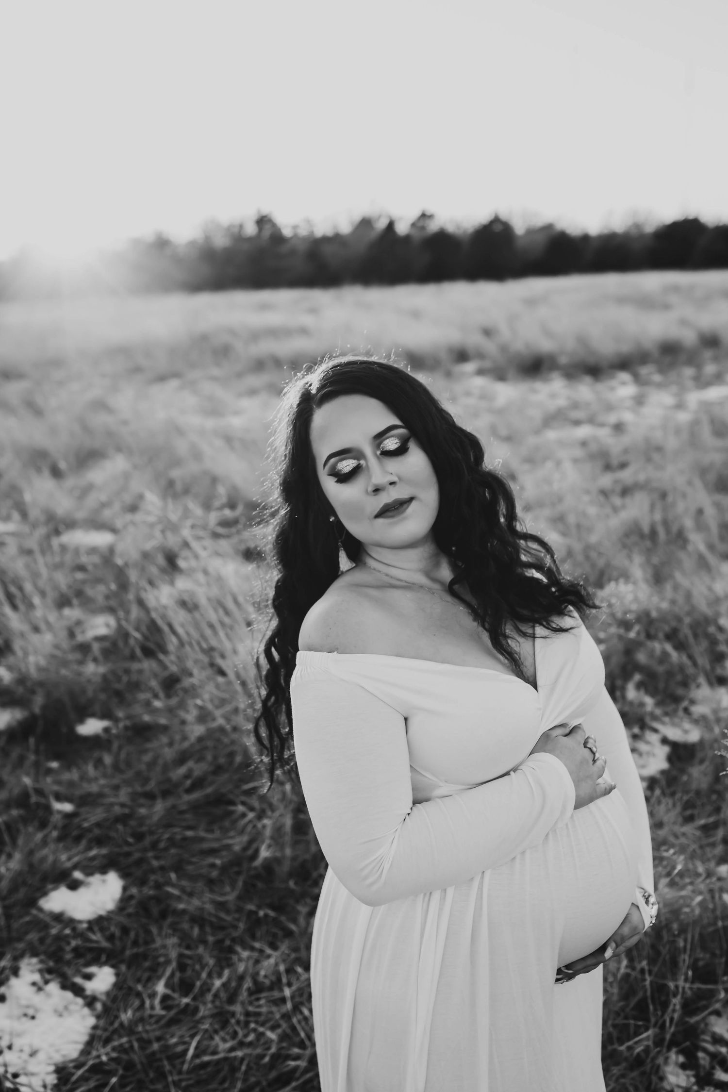 Black and white maternity photo of a pregnant woman in a white off-shoulder gown standing in an open field.