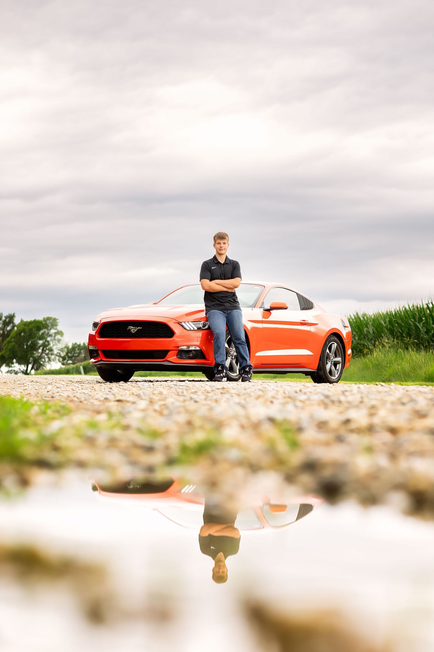 Young man with crossed arms leaning against a red Ford Mustang on a gravel road, reflected in a puddle below.