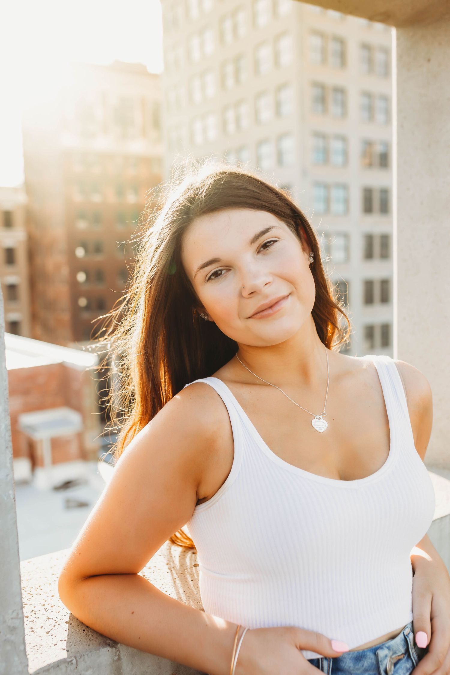 Young woman in white tank top smiling on urban rooftop at golden hour with city buildings in background.