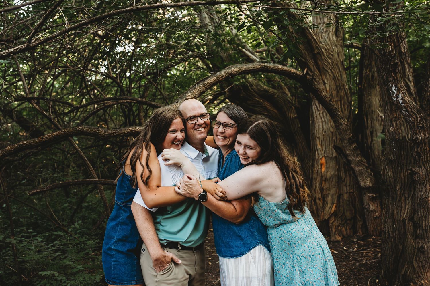 A joyful family of four hugging and laughing together outdoors among lush green trees.