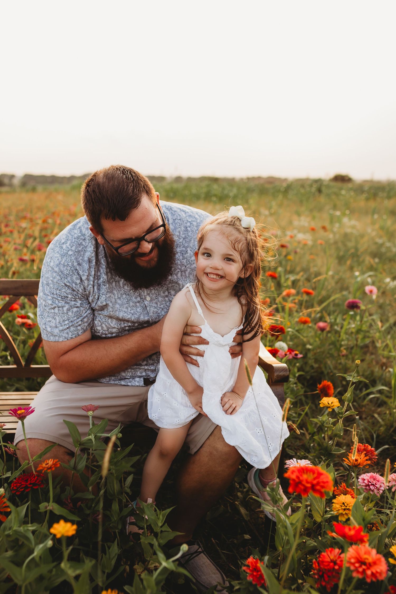A bearded father with glasses laughs with his young daughter in a white dress among colorful flowers in a field.