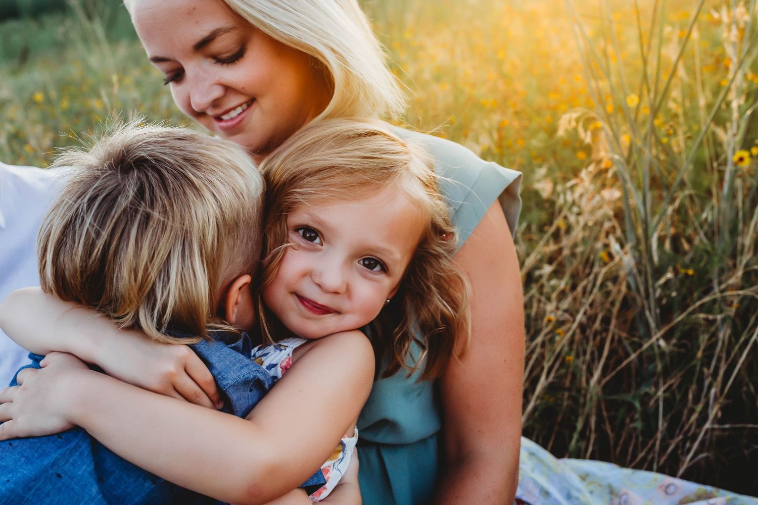 Mother tenderly embracing two young children in a sunlit meadow during golden hour.