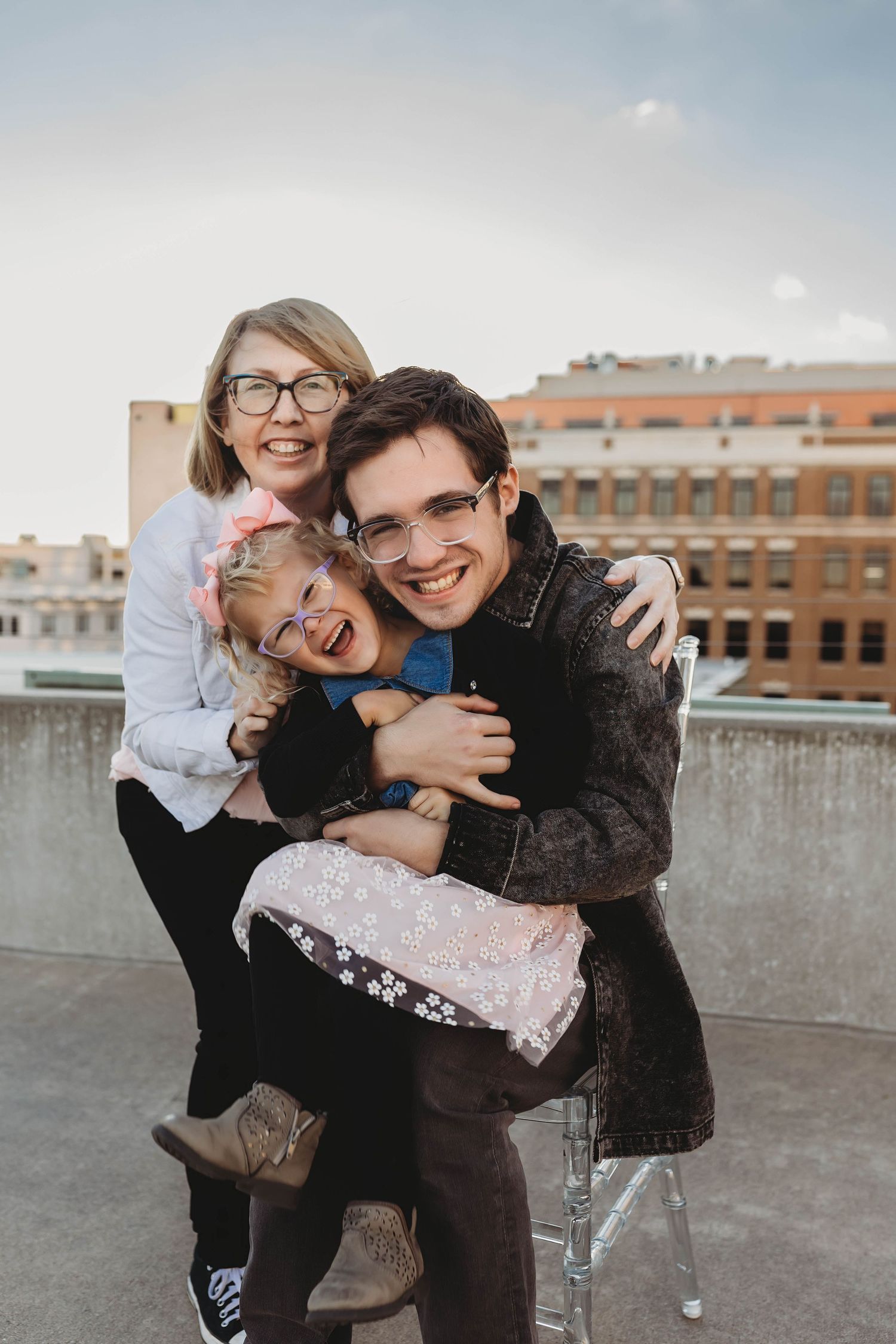 Family of three smiling and hugging on a rooftop, with a young girl in pink between two adults wearing glasses.