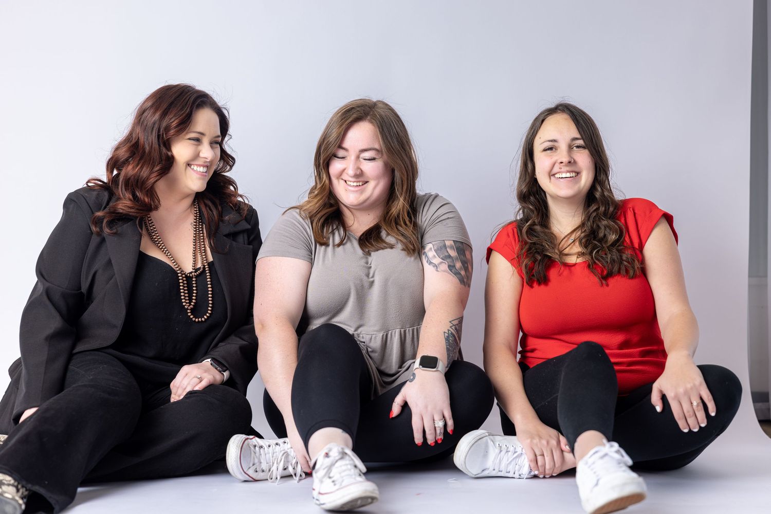 Three friends sitting together against gray wall wearing casual outfits.