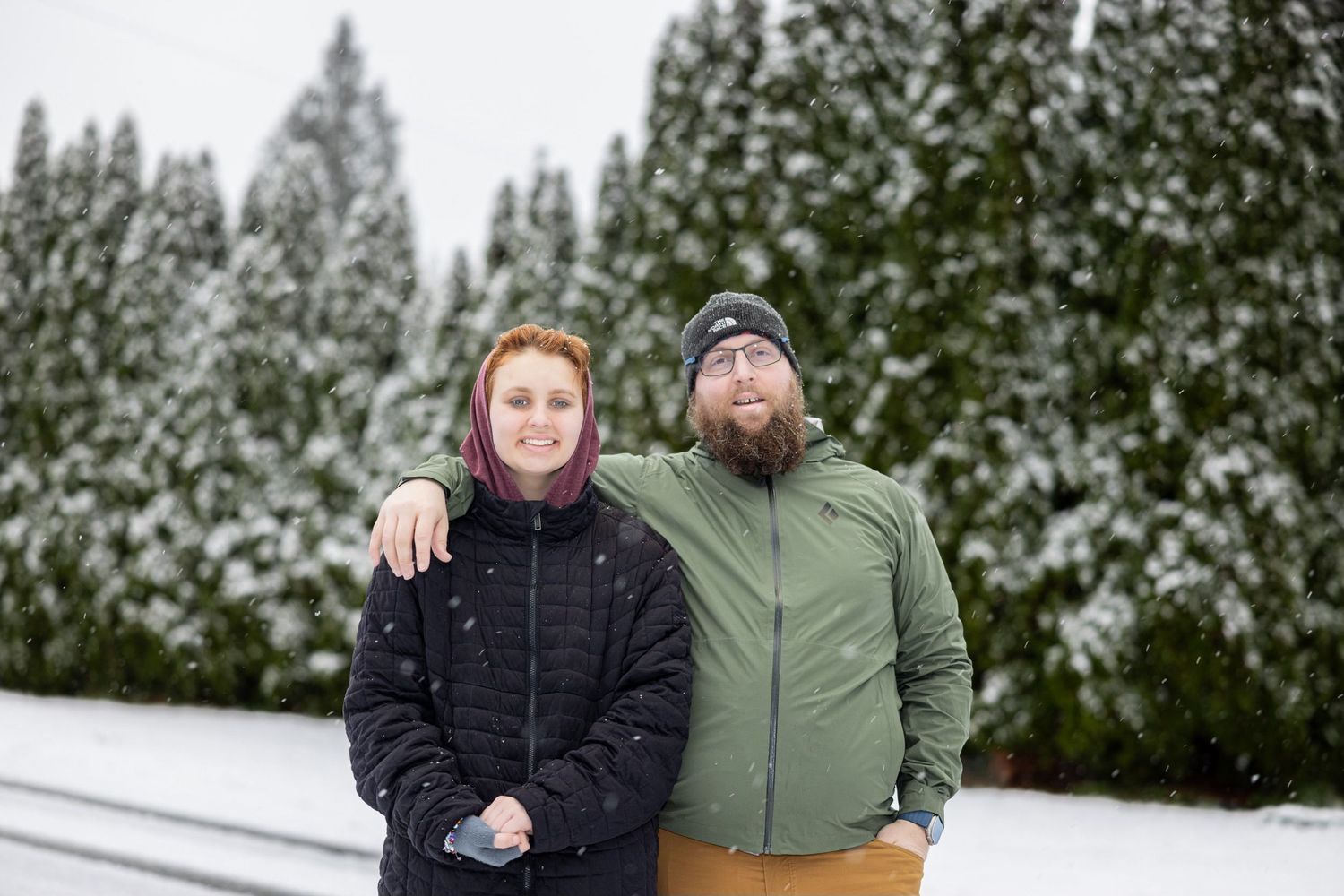 Two people standing together in winter coats with snow-covered trees in background.