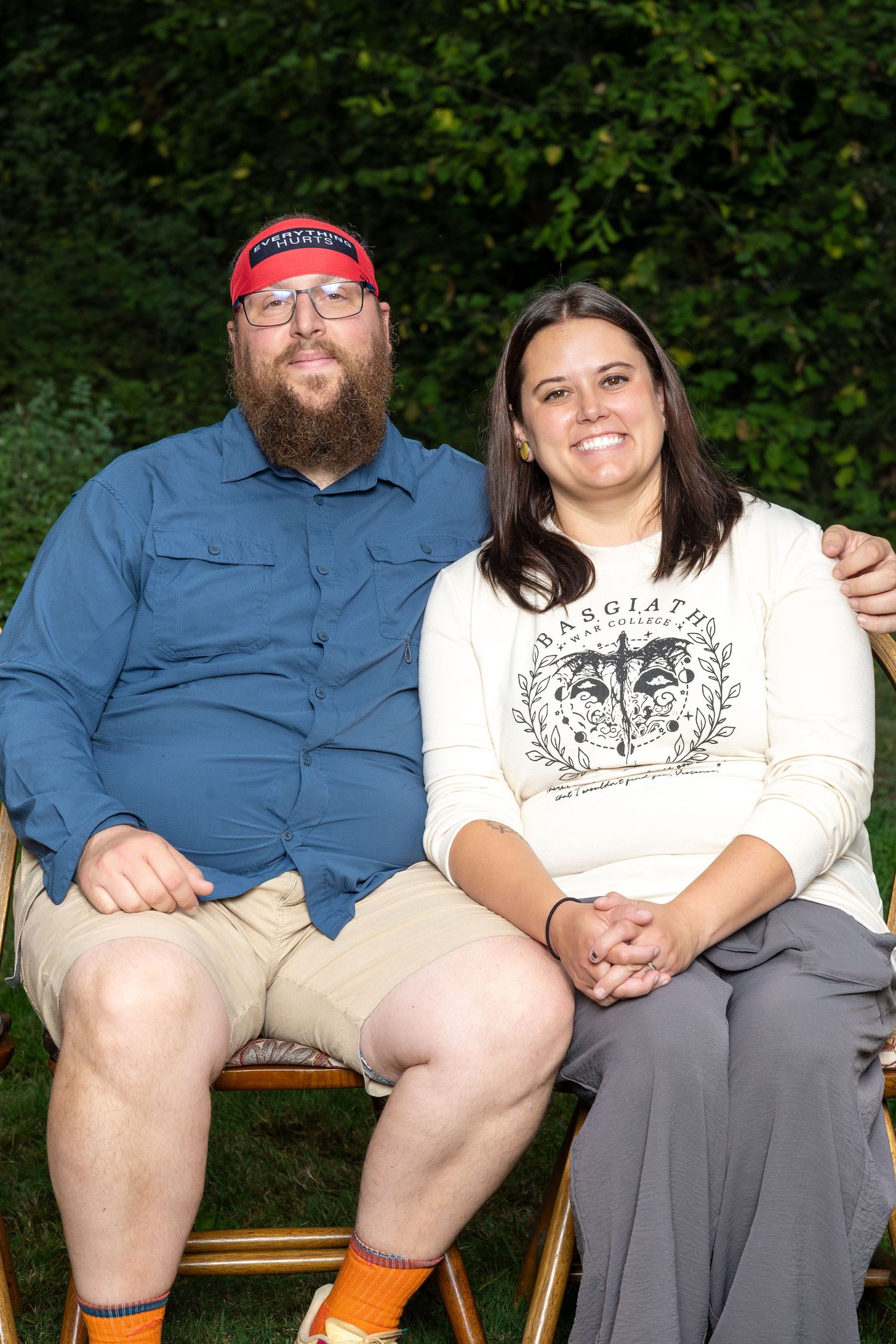 A couple sits together outdoors on wooden chairs with greenery in the background.