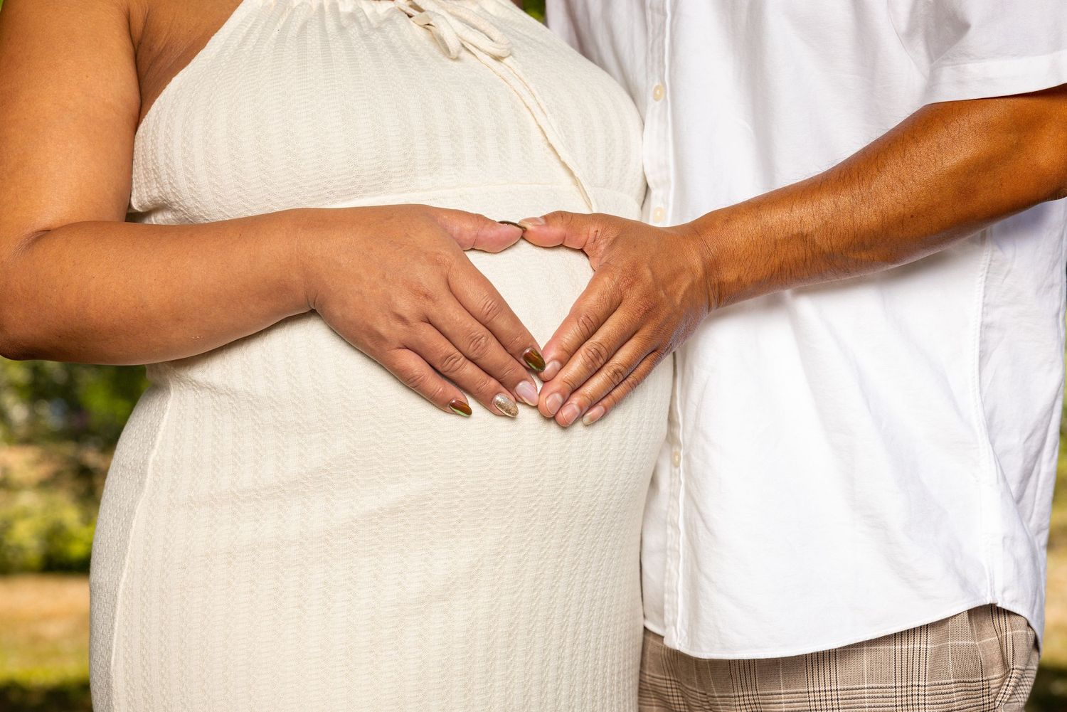 Close-up of hands forming heart shape on pregnant belly in white dress outdoors.