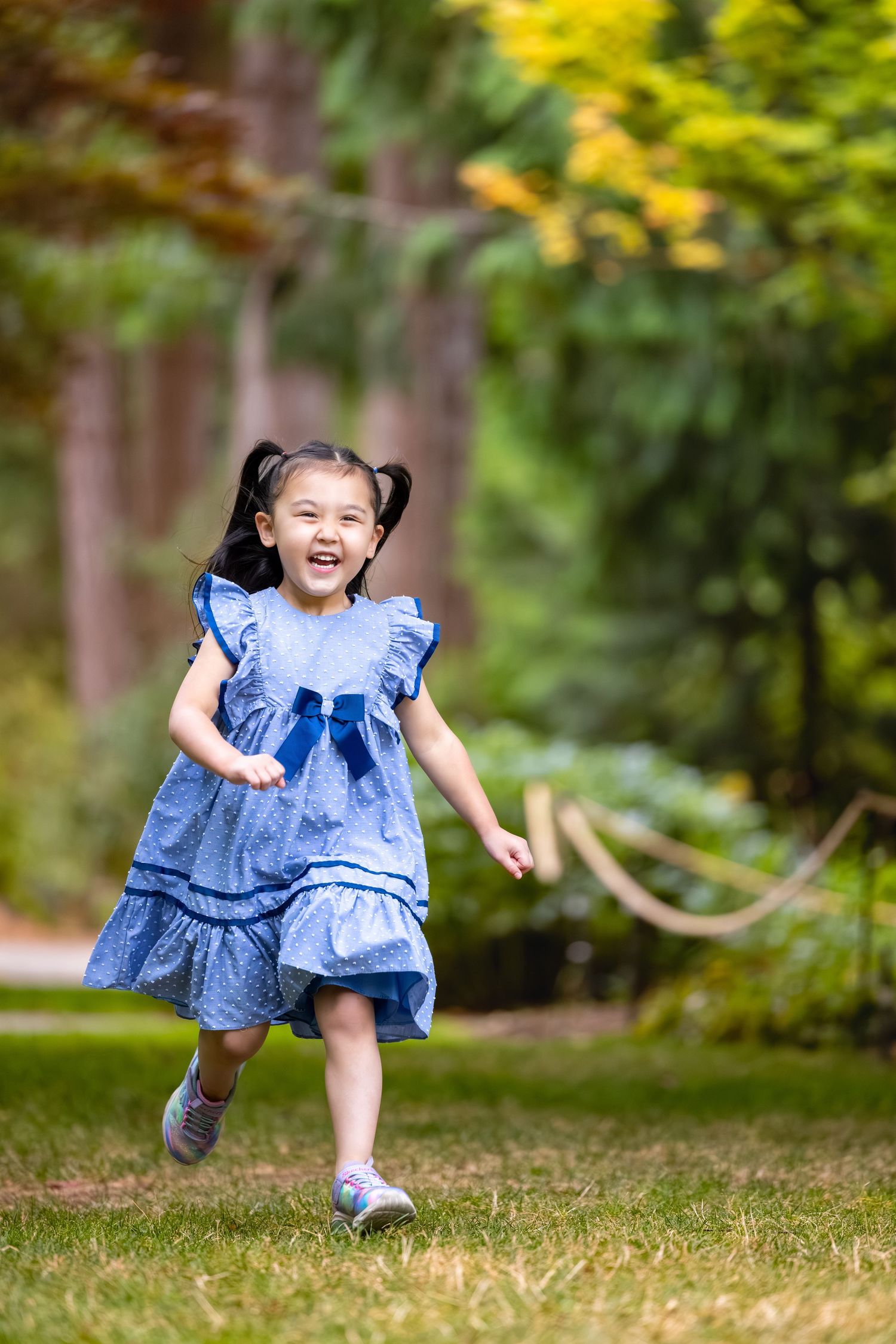 A young girl in a blue ruffled dress runs joyfully through a park with yellow flowers in background.