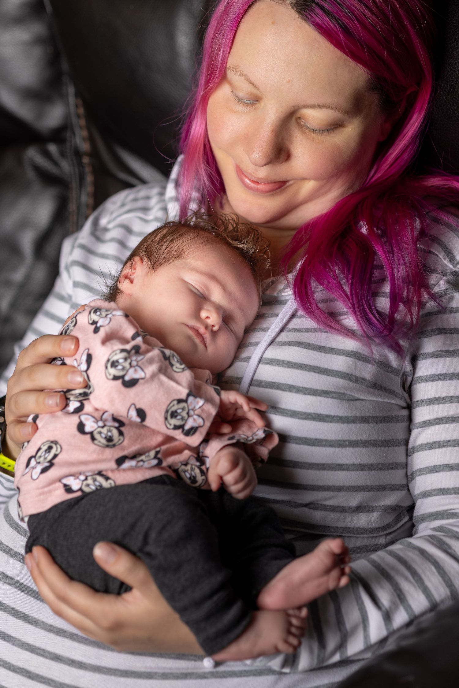 Parent with bright pink hair holding sleeping infant in striped outfit.