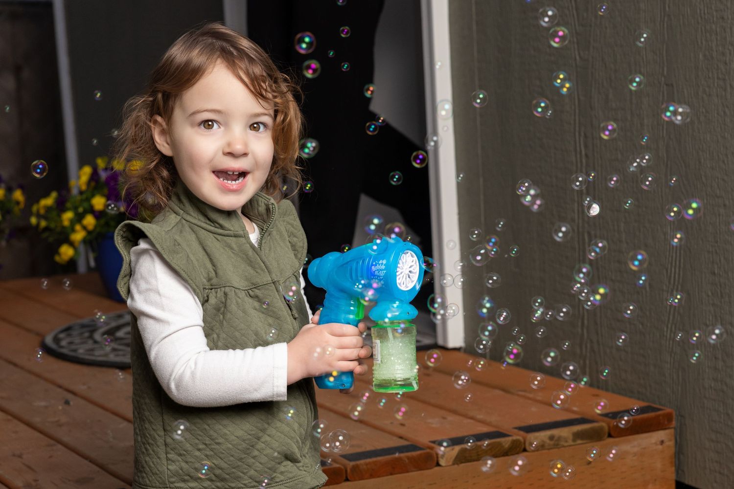 Child in green vest playing with blue bubble gun on wooden deck with bubbles floating in air.