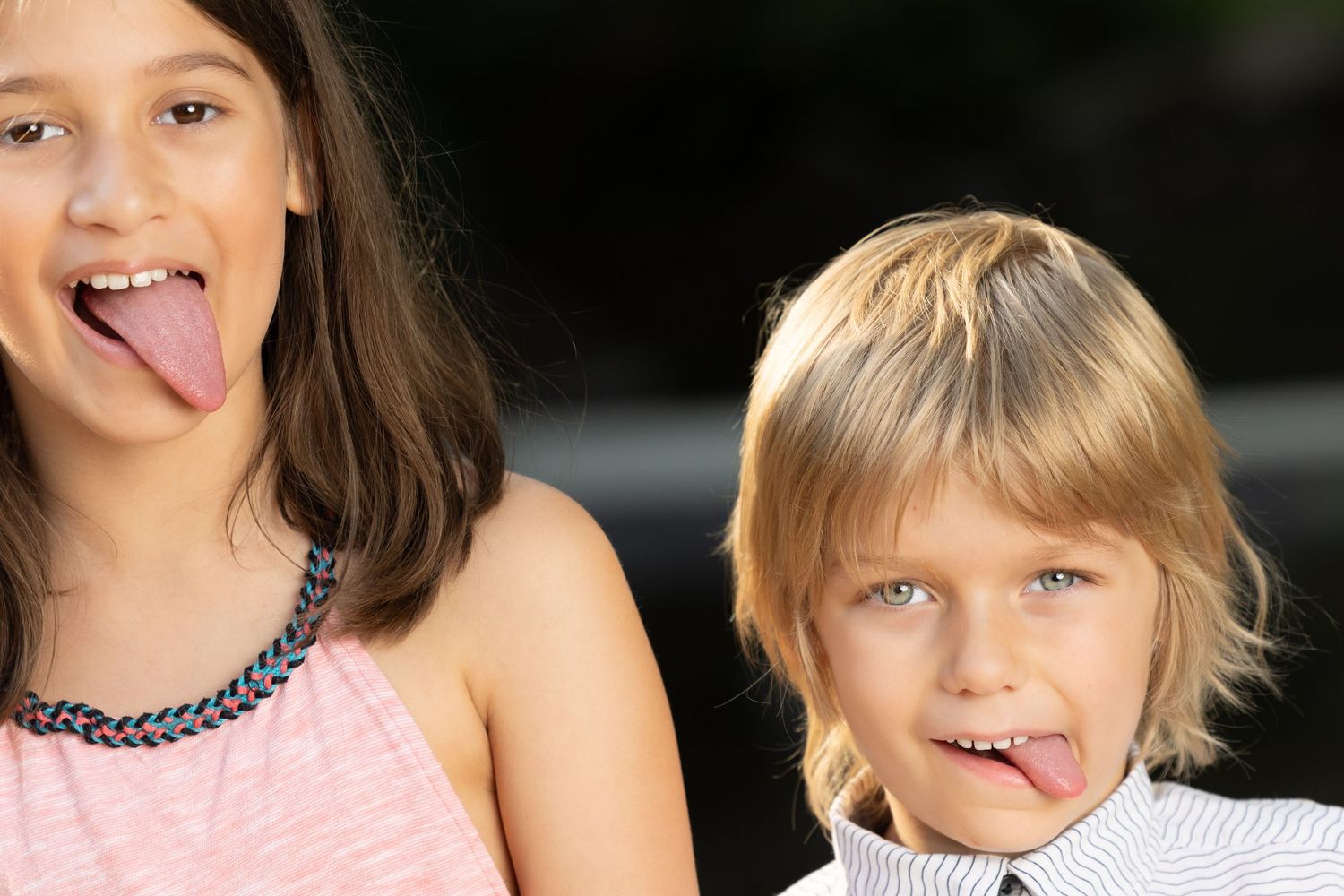 Two playful children sticking out tongues while posing together outdoors.