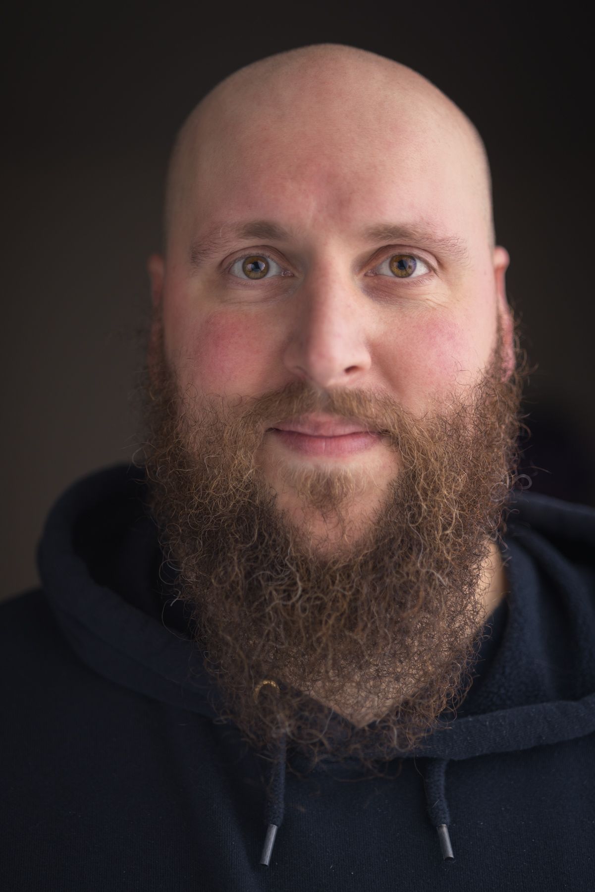 Portrait of a bald man with a full beard wearing a dark hoodie against black background.